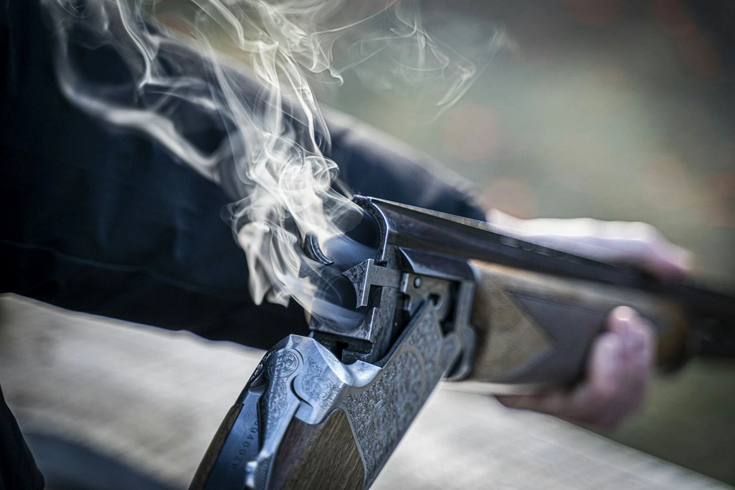 A person is visible repairing or assembling a metal firearm, with visible details like engraved patterns, in an outdoor setting. Smoke or steam rises from the gun's trigger or barrel area.