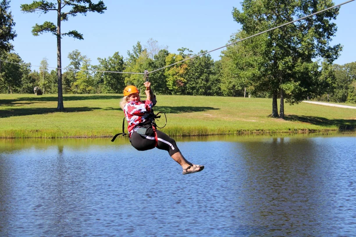Person ziplining over a lake on a sunny day, wearing a safety helmet and harness.