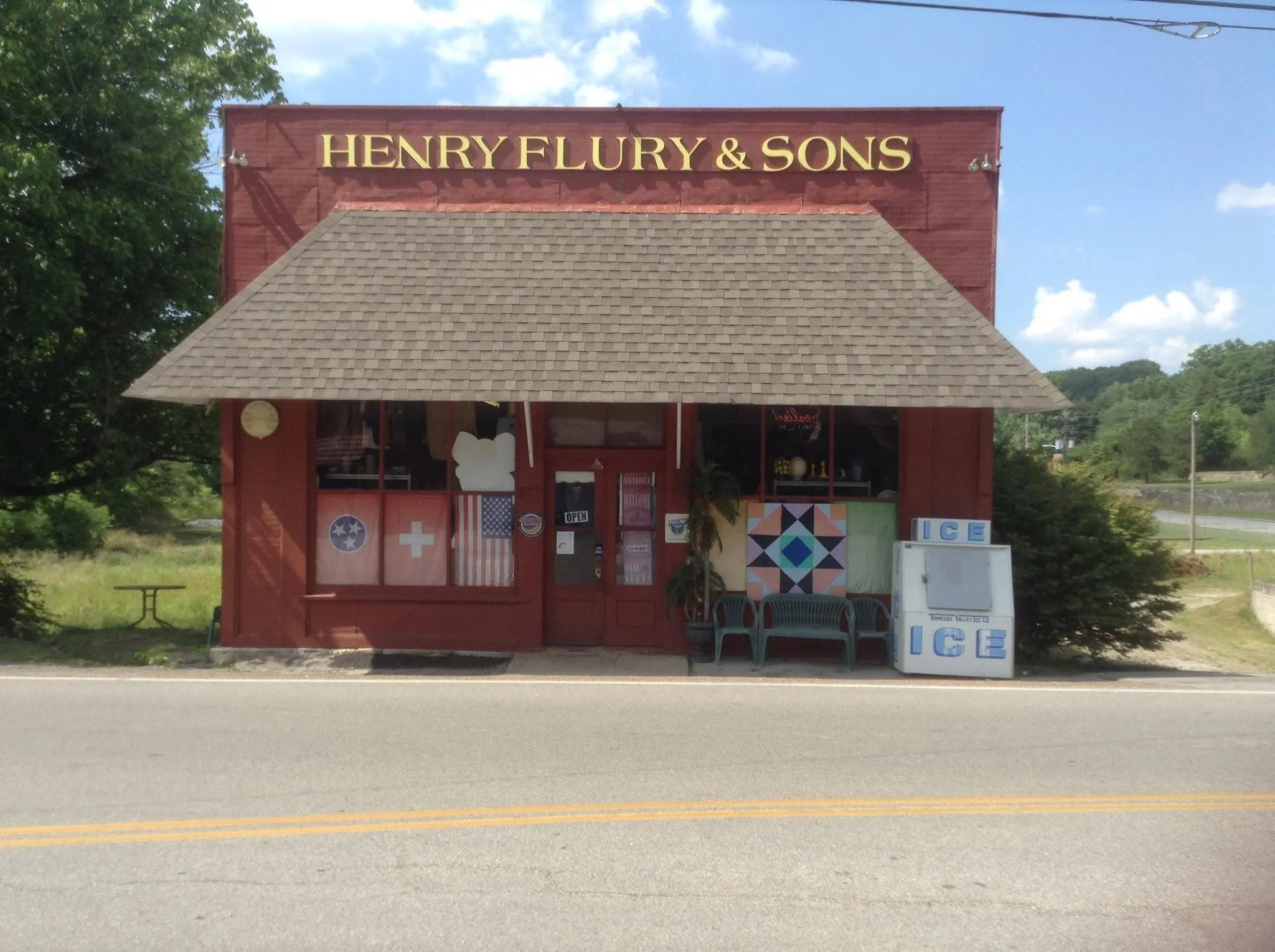A small wooden building with a shingled roof, featuring a sign that reads 'Henry Flury & Sons'. The building is decorated with patriotic symbols like flags, and has a row of chairs outside. There is a white ice machine next to the chairs. The background includes trees, a partly cloudy sky, and power lines.