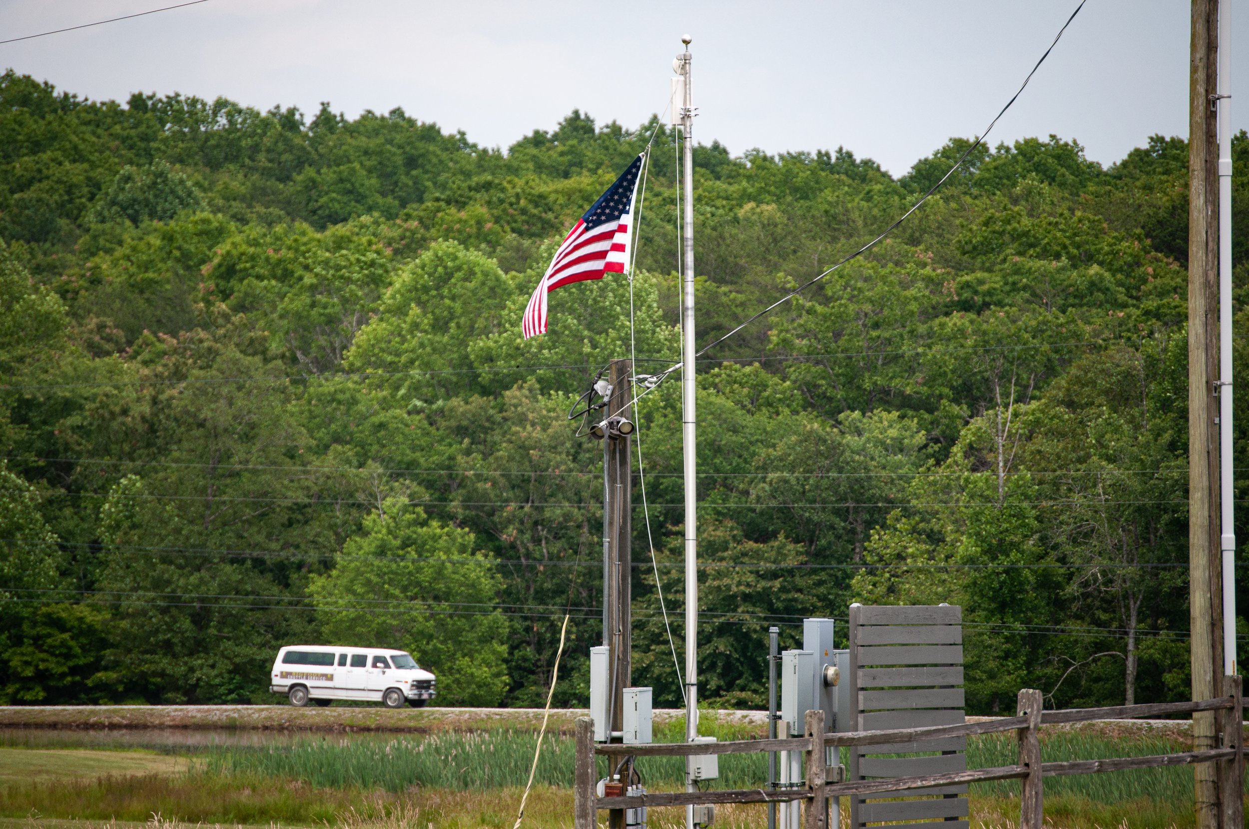 American flag flying on a flagpole with a green, wooded hillside in the background. There is a white van on a road and a wooden fence in the foreground.