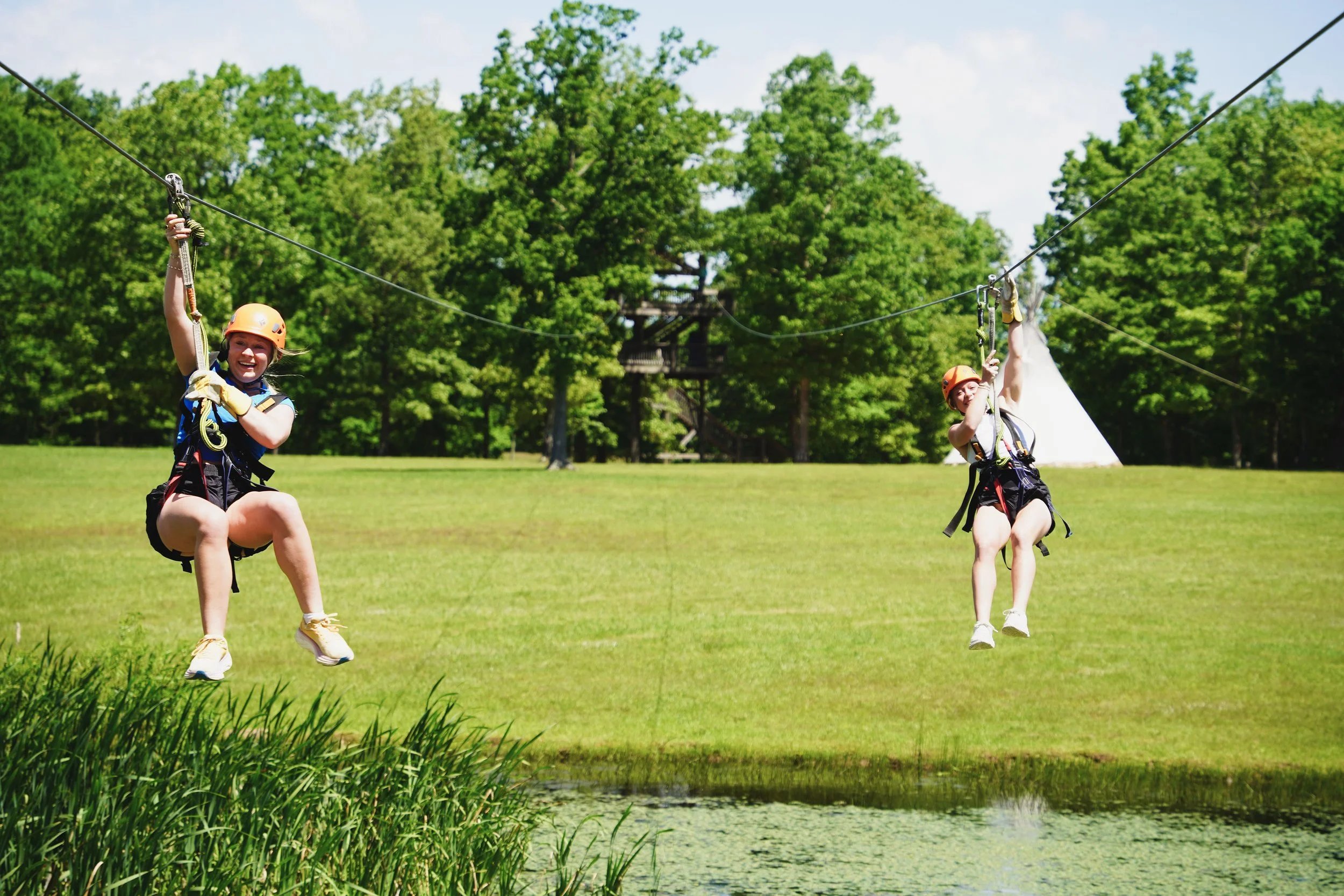 Two children wearing helmets and harnesses ziplining over a pond in a green outdoor area.