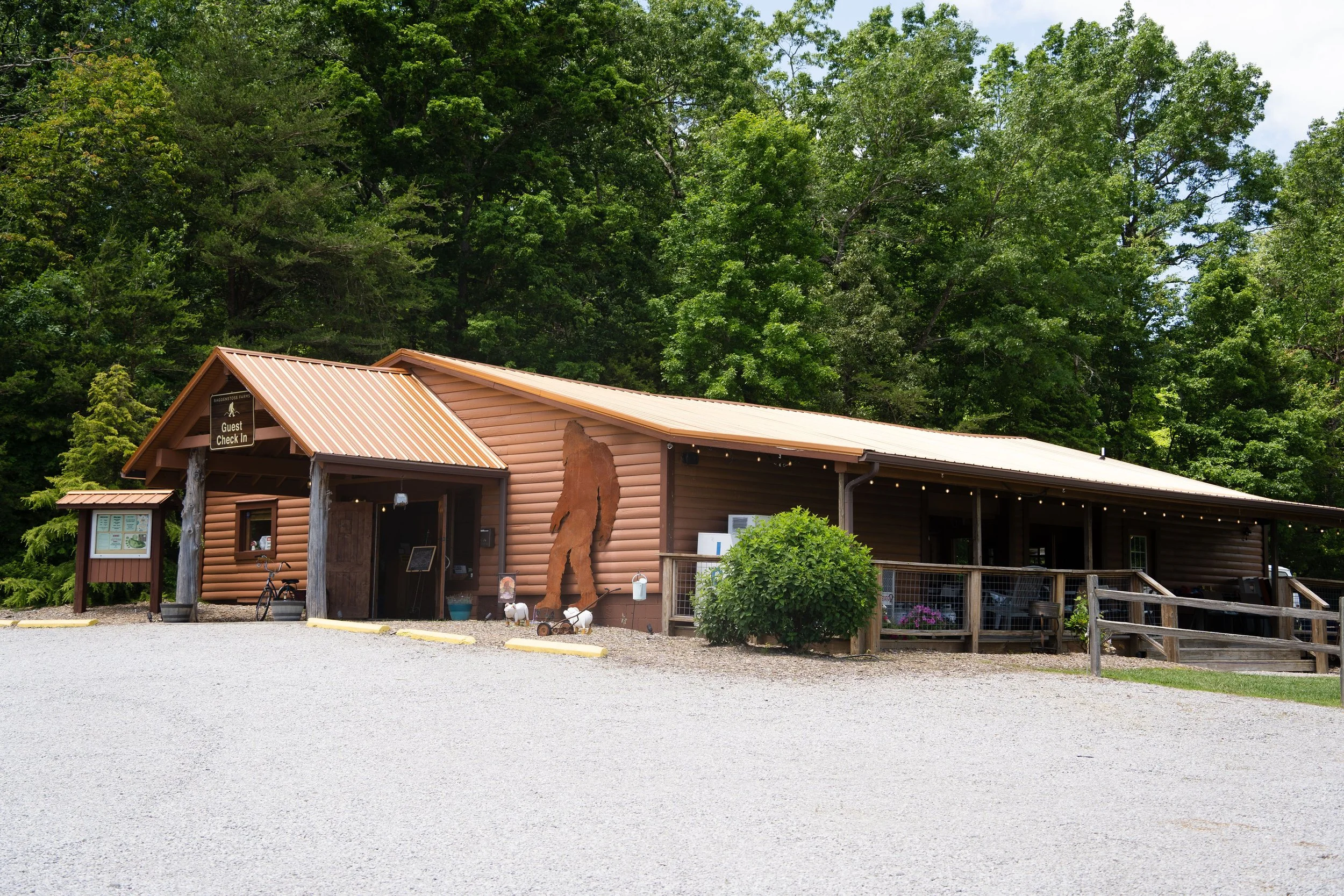A rustic visitor center building with a log cabin style, featuring a metal roof, located in a lush, green, wooded area. There is a sign that says "Guest Check In" and decorative items such as a wooden bear silhouette and sheep figures outside.
