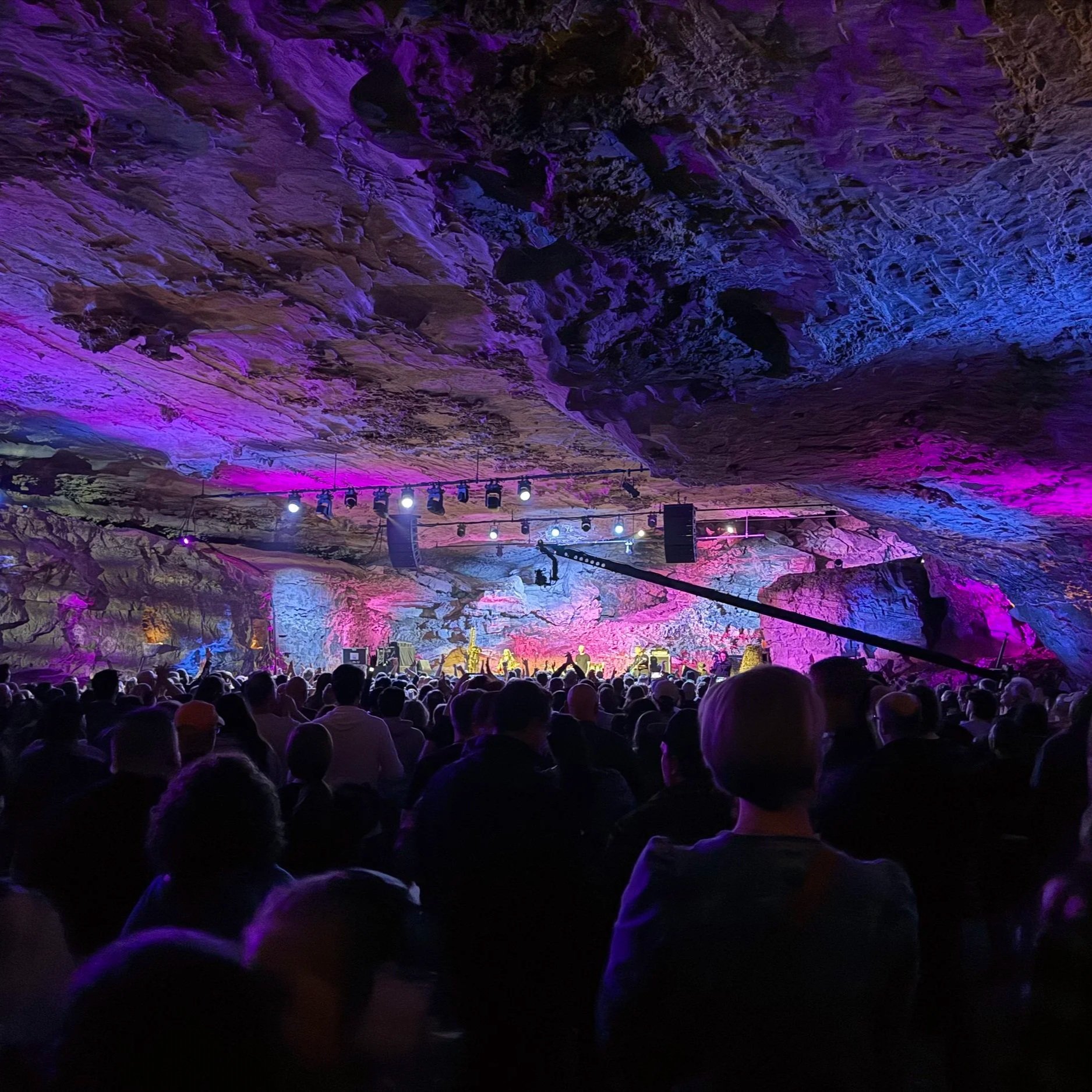 A large crowd of people gathered inside a cavern with rock formations illuminated by purple, pink, and blue lighting. There is a stage at the front with performers and speakers, and various stage equipment and lighting fixtures are visible.