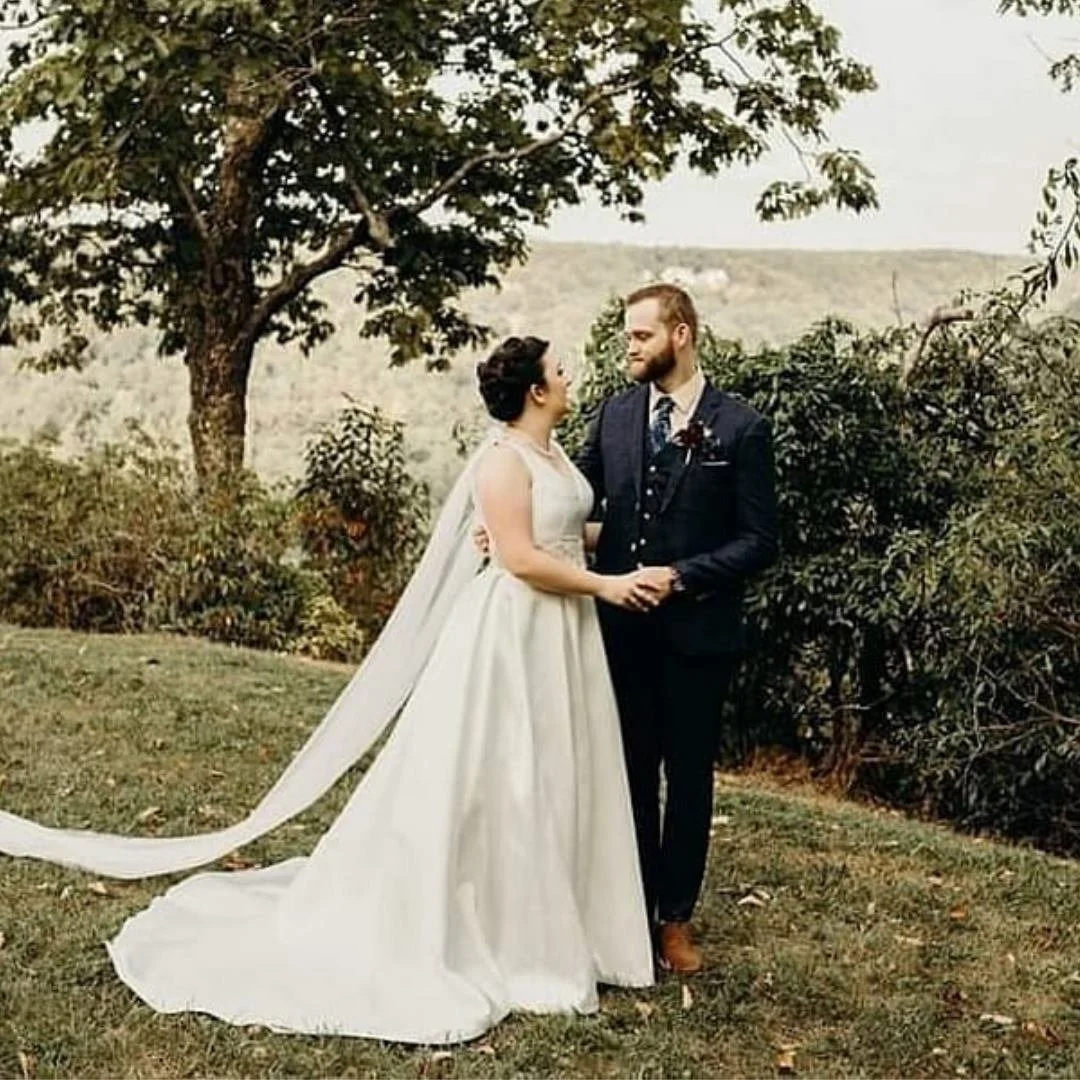 A bride and groom holding hands and looking at each other outdoors on their wedding day, with trees and a scenic landscape in the background.