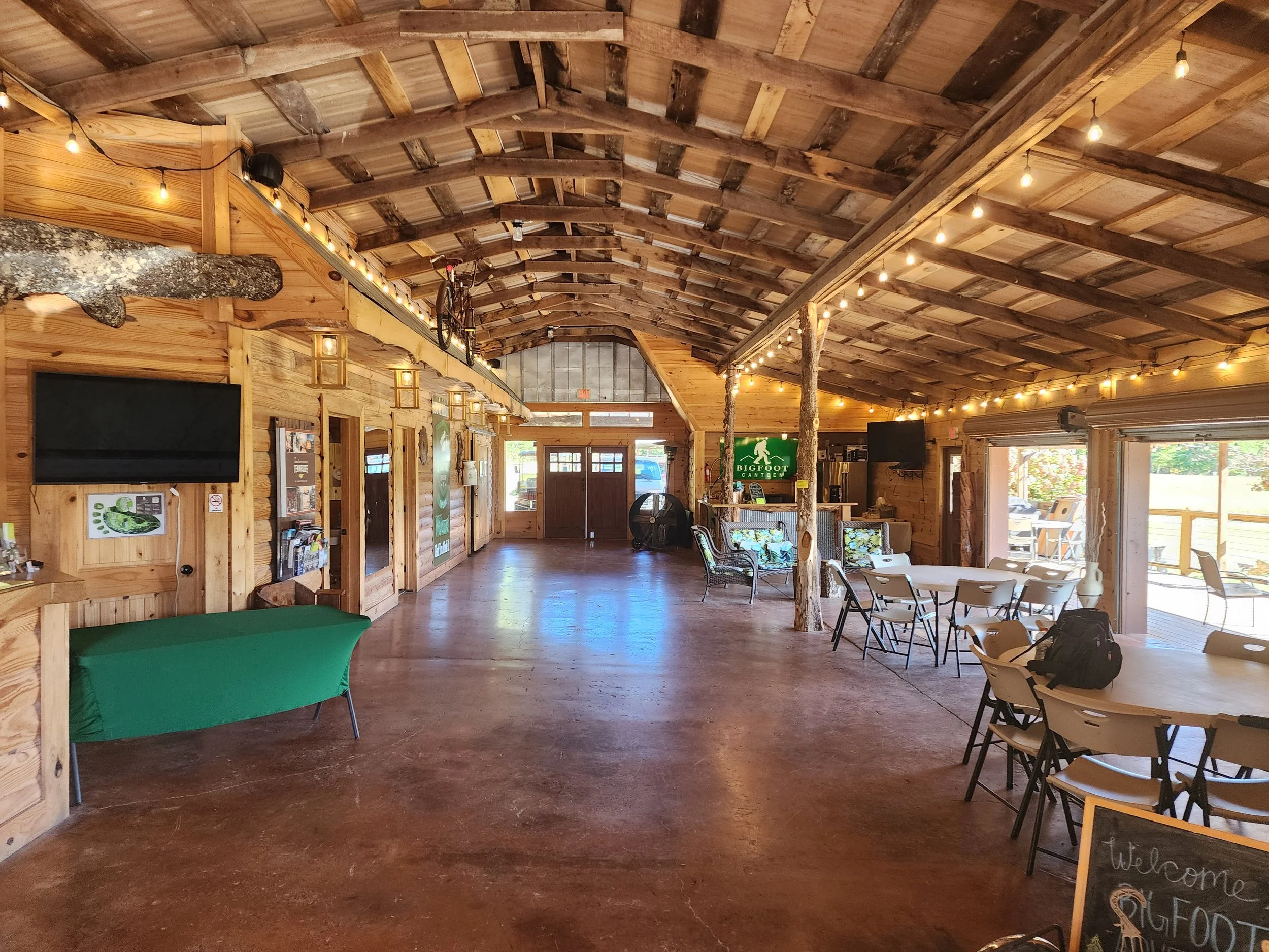 Interior of a rustic wooden building with string lights hanging from the ceiling, a wooden wall with framed pictures, a television, a green table, and a seating area with chairs and tables near large windows.