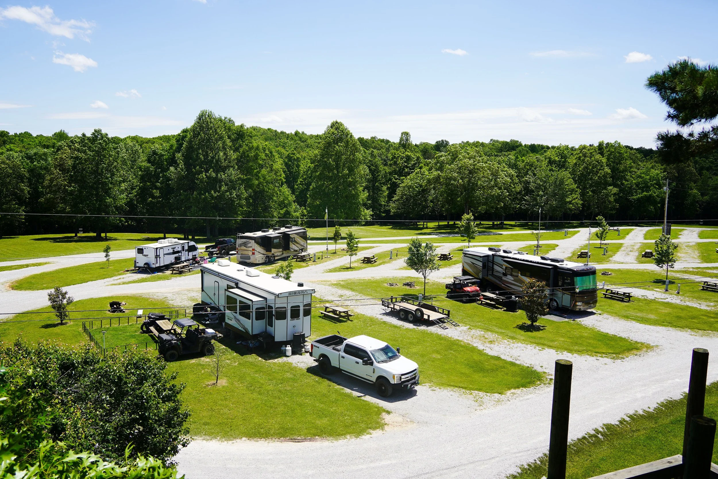 A campground with multiple RVs, trucks, picnic tables, and trees, under a blue sky with scattered clouds.