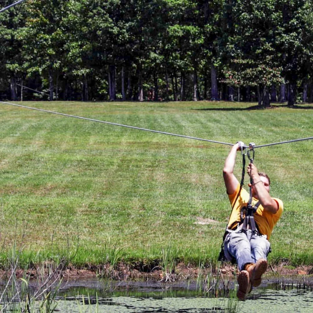 Person wearing a yellow shirt and gray shorts ziplining over a pond in a grassy park with trees in the background.