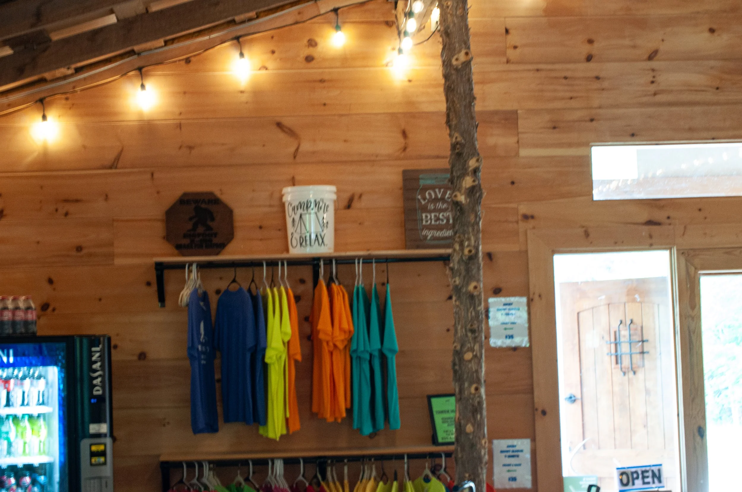 Wood-paneled interior with string lights, colorful shirts hanging on a rack, a vending machine on the left, and a wooden door on the right. Decor includes signs and buckets on a shelf.