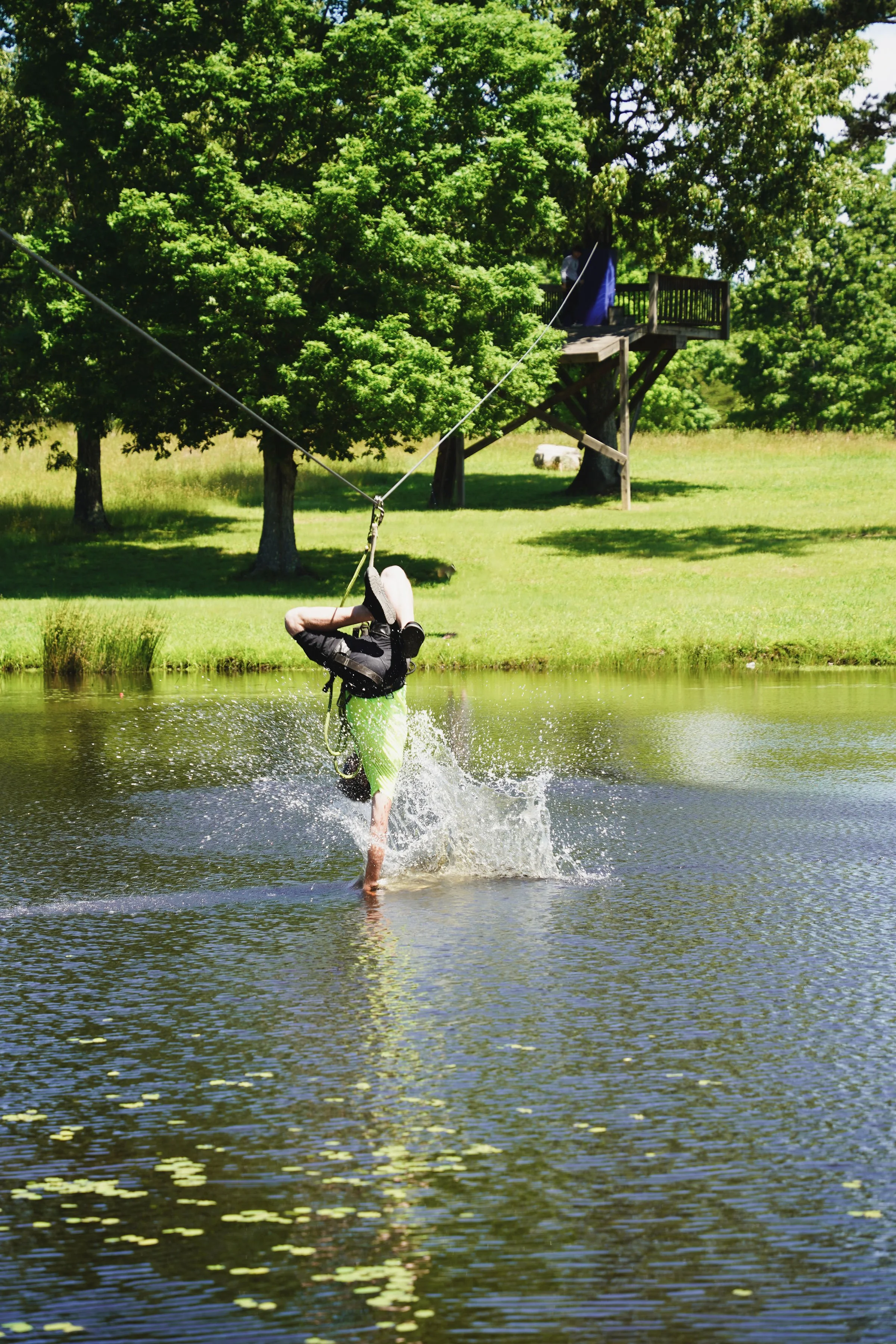 Person in a bright green shirt and dark shorts performing a handstand on a wakeboard in the water, with a treehouse and lush trees in the background.