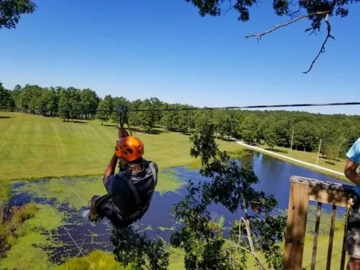 A child wearing a helmet and harness is zip-lining over a lake and grassy field on a sunny day with a clear blue sky.