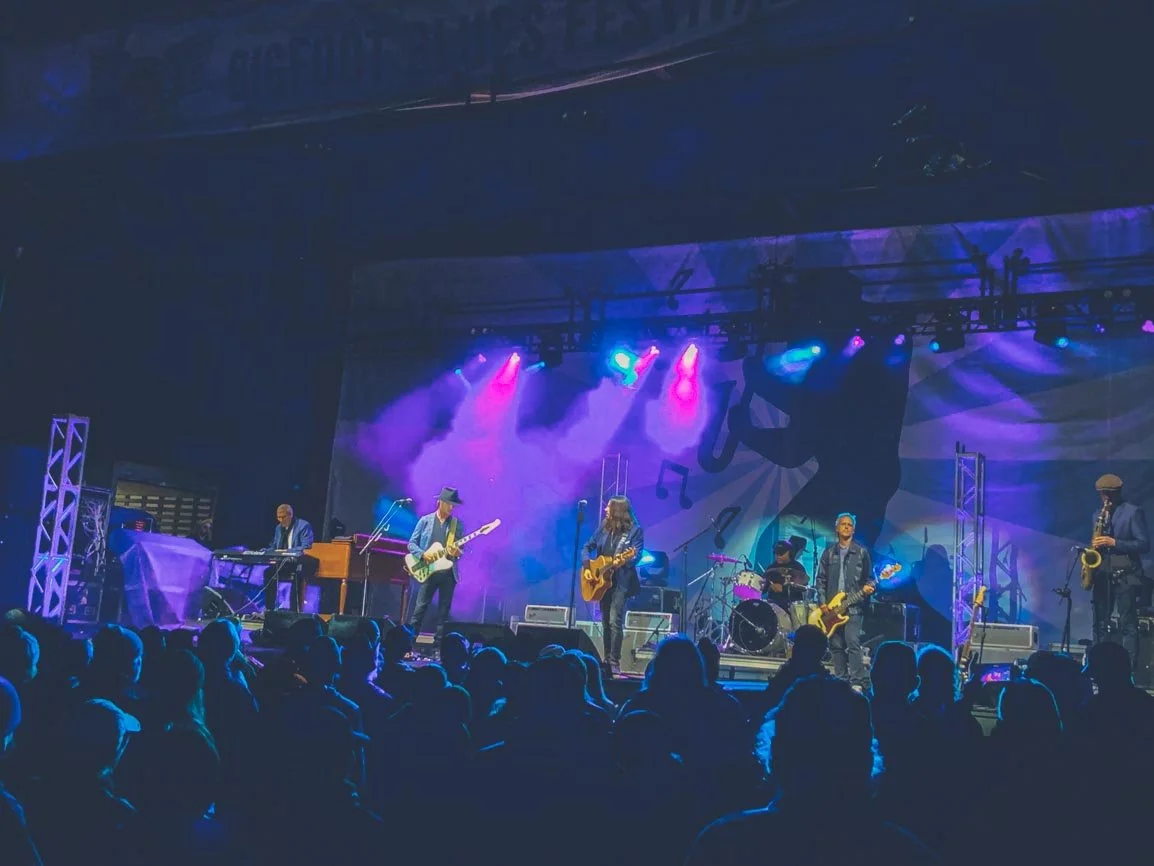 A band of six musicians performing on stage with colorful purple and blue lighting, audience in foreground, backdrop featuring a silhouette of a musician and musical notes.