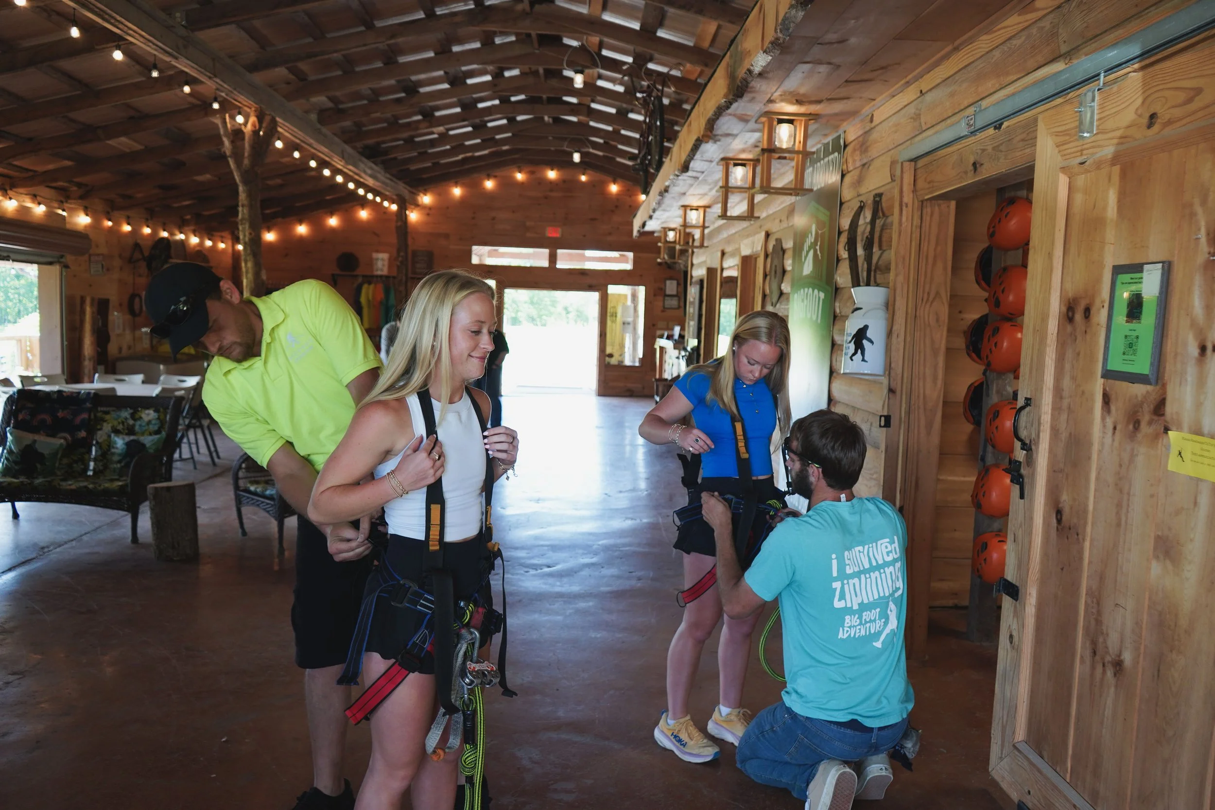 Two women are receiving instruction from a guide about ziplining safety gear inside a wooden lodge. One man is helping a woman put on her harness. The scene is set in a rustic indoor area with wood walls and ceiling, string lights, and outdoor light 
