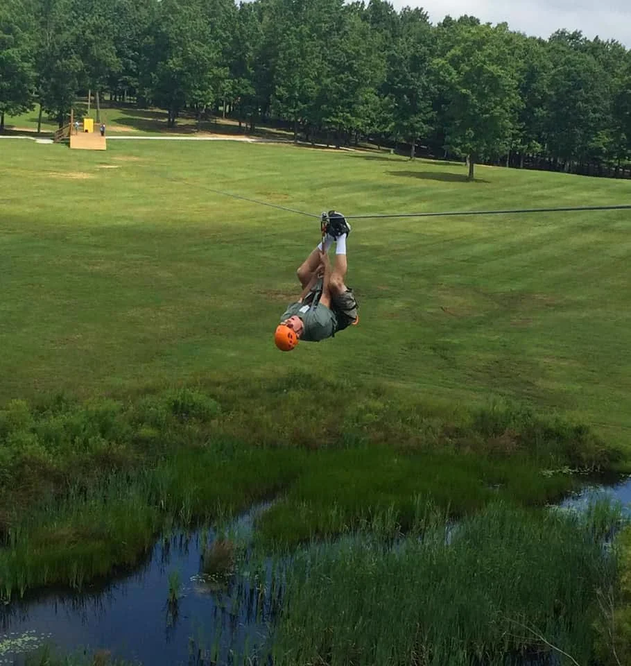 Person zip-lining over a grassy field next to a small stream, with trees in the background.