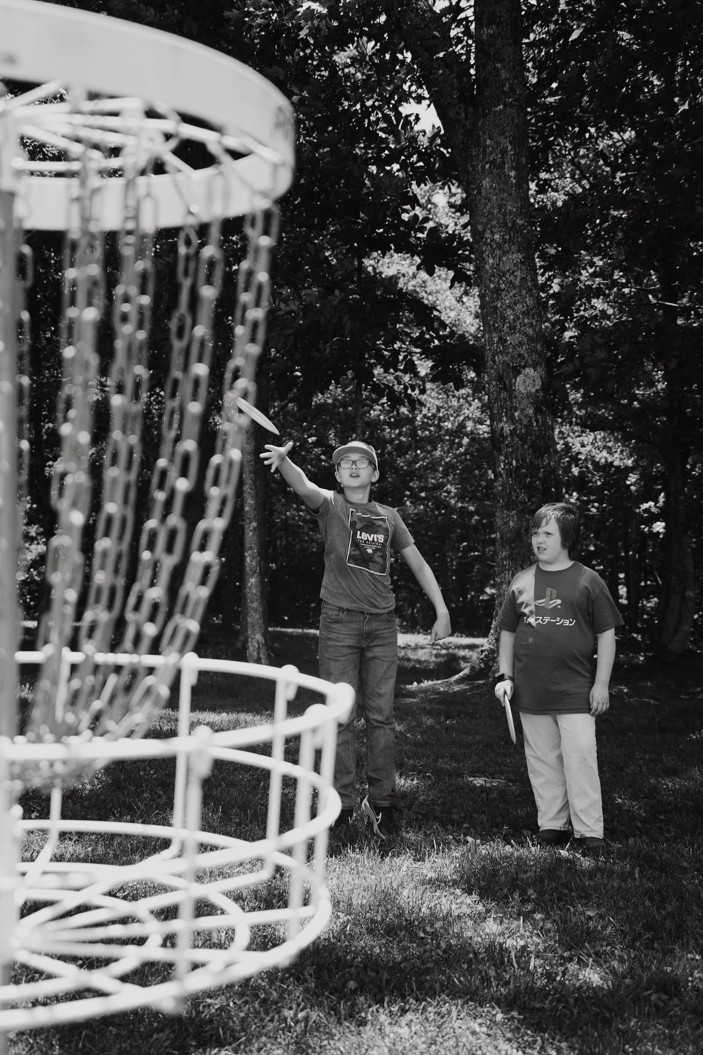 Two boys playing disc golf outdoors, one throwing a disc towards a metal disc golf basket, surrounded by trees and grass.