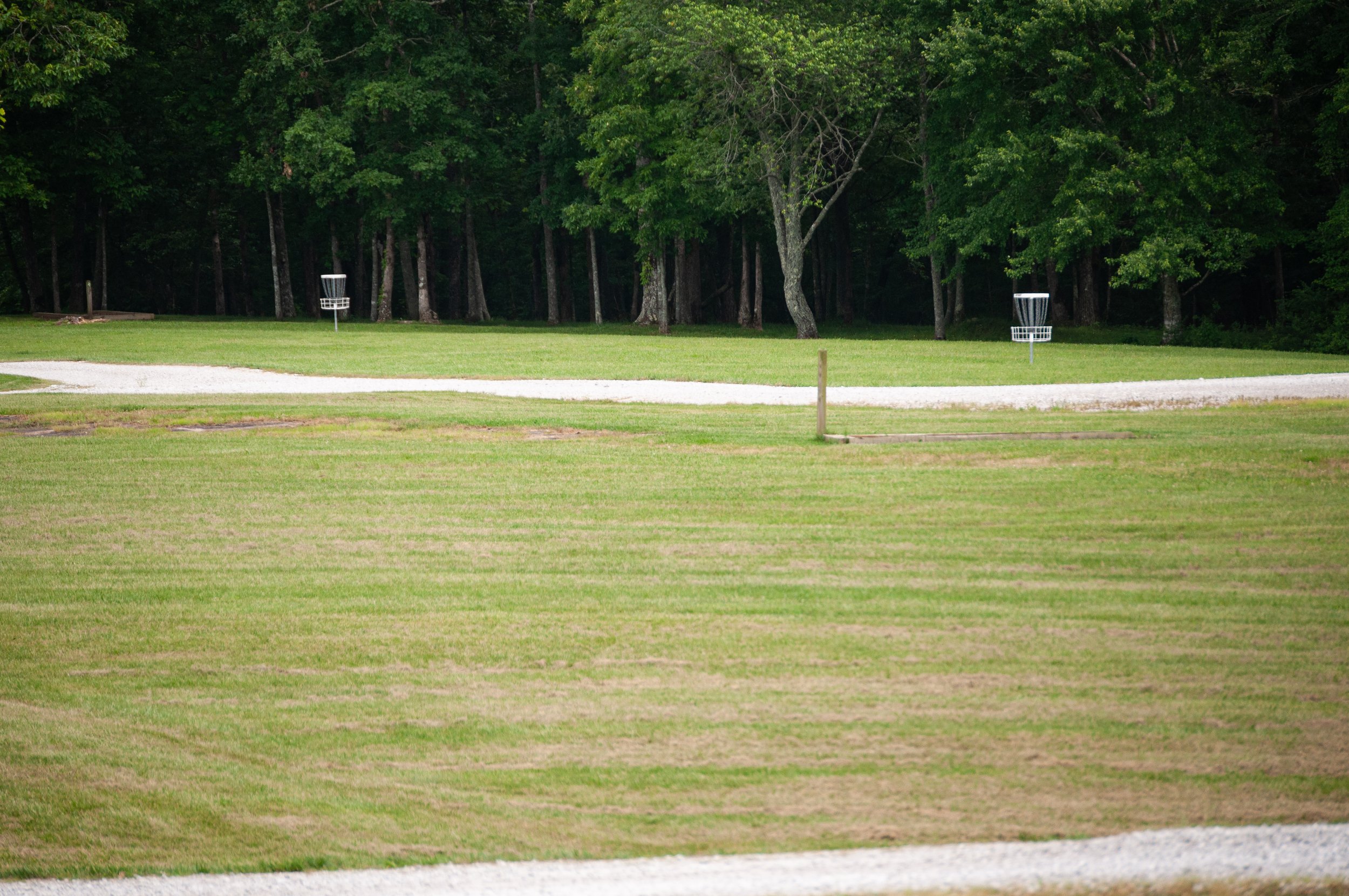 A disc golf course with two baskets and a gravel cart path, surrounded by green grass and trees in the background.