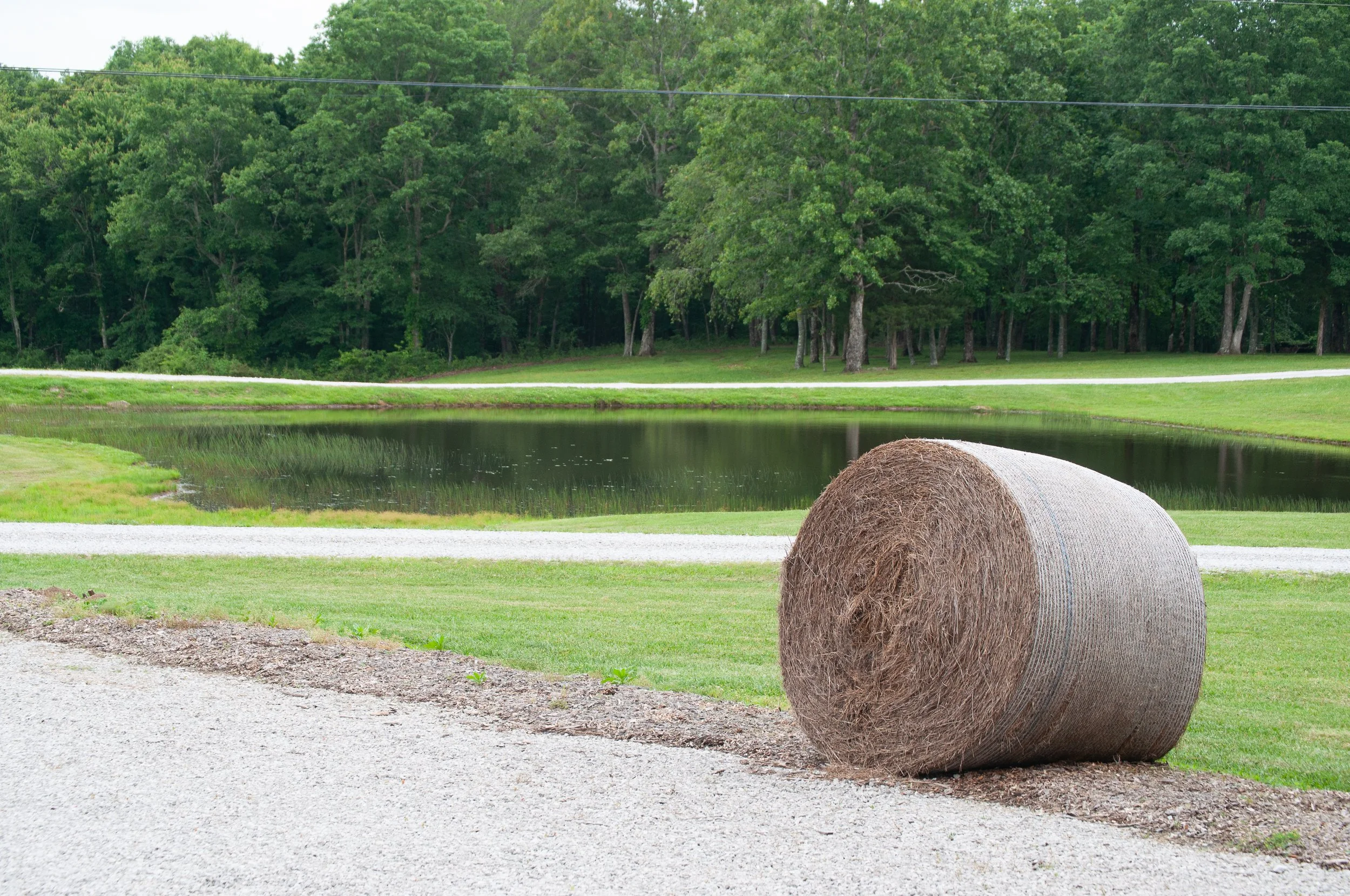 Round hay bale resting near a gravel path with a peaceful fishing pond and green forest backdrop at Bigfoot Adventure in Tracy City, Tennessee.