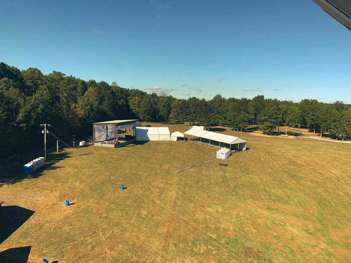 Large outdoor event setup with a stage and multiple tents on a grassy field, surrounded by trees under a clear sky.