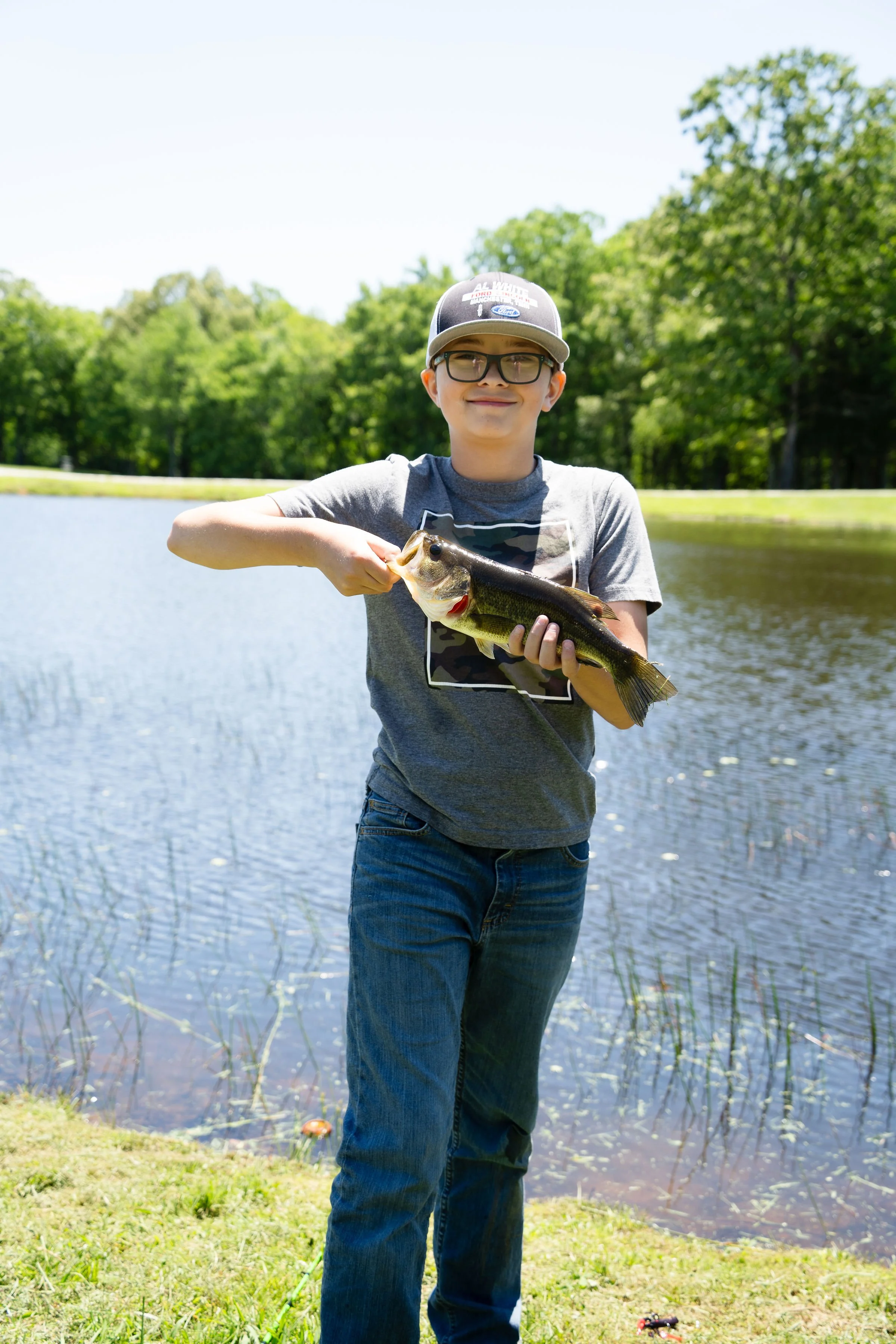 Young boy holding a fish by a pond with trees in the background on a sunny day.