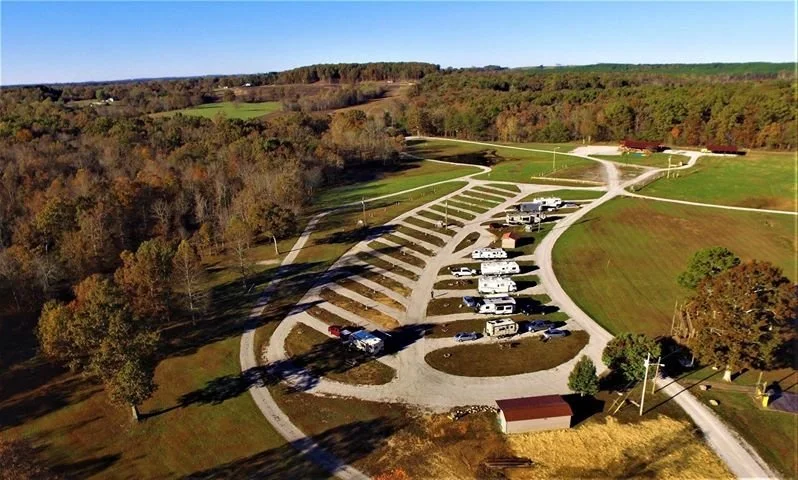 Aerial view of a campground with RVs and trailers parked along curved gravel roads, surrounded by green fields and trees with a forested hillside in the background.