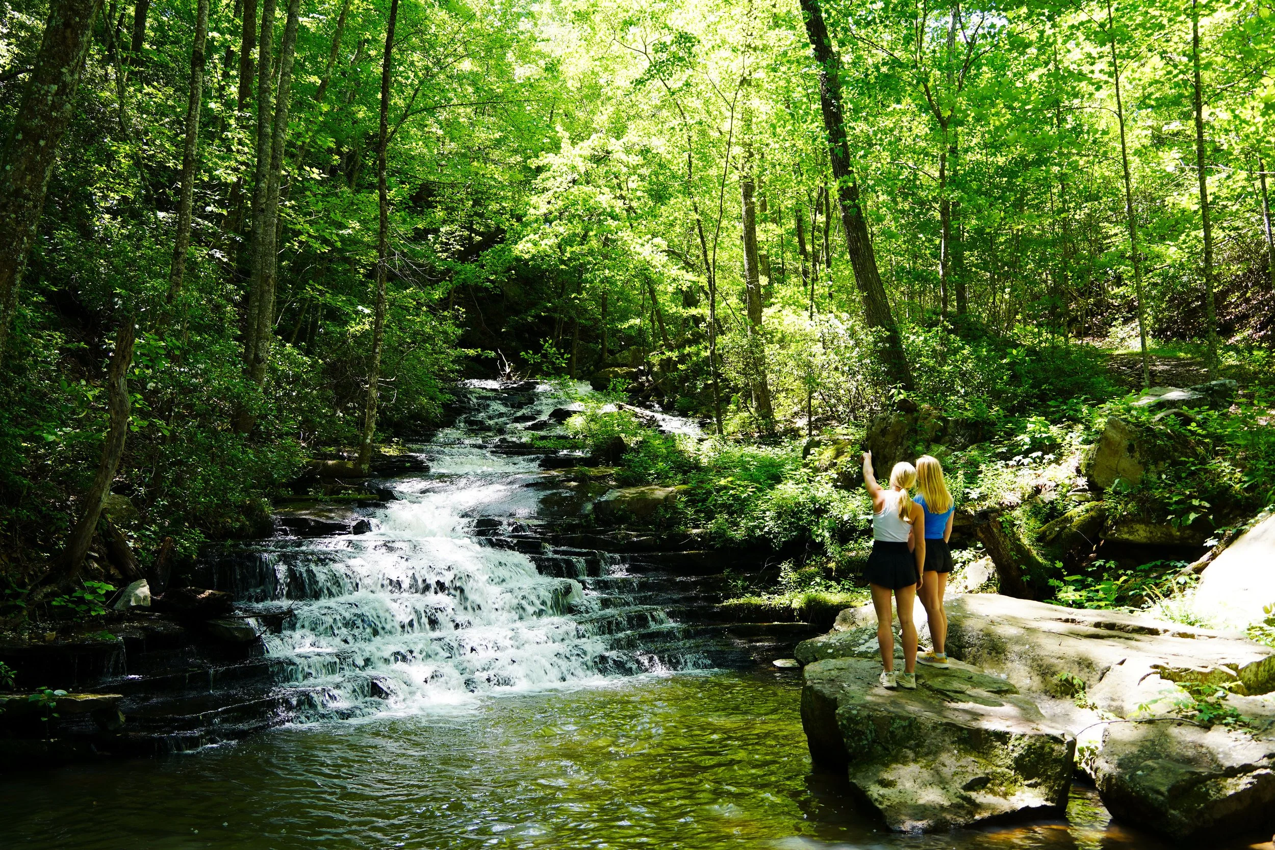 Two blonde women in summer clothing stand on a large rock by a forest stream, taking a selfie amidst lush green trees and sunlight filtering through the leaves.