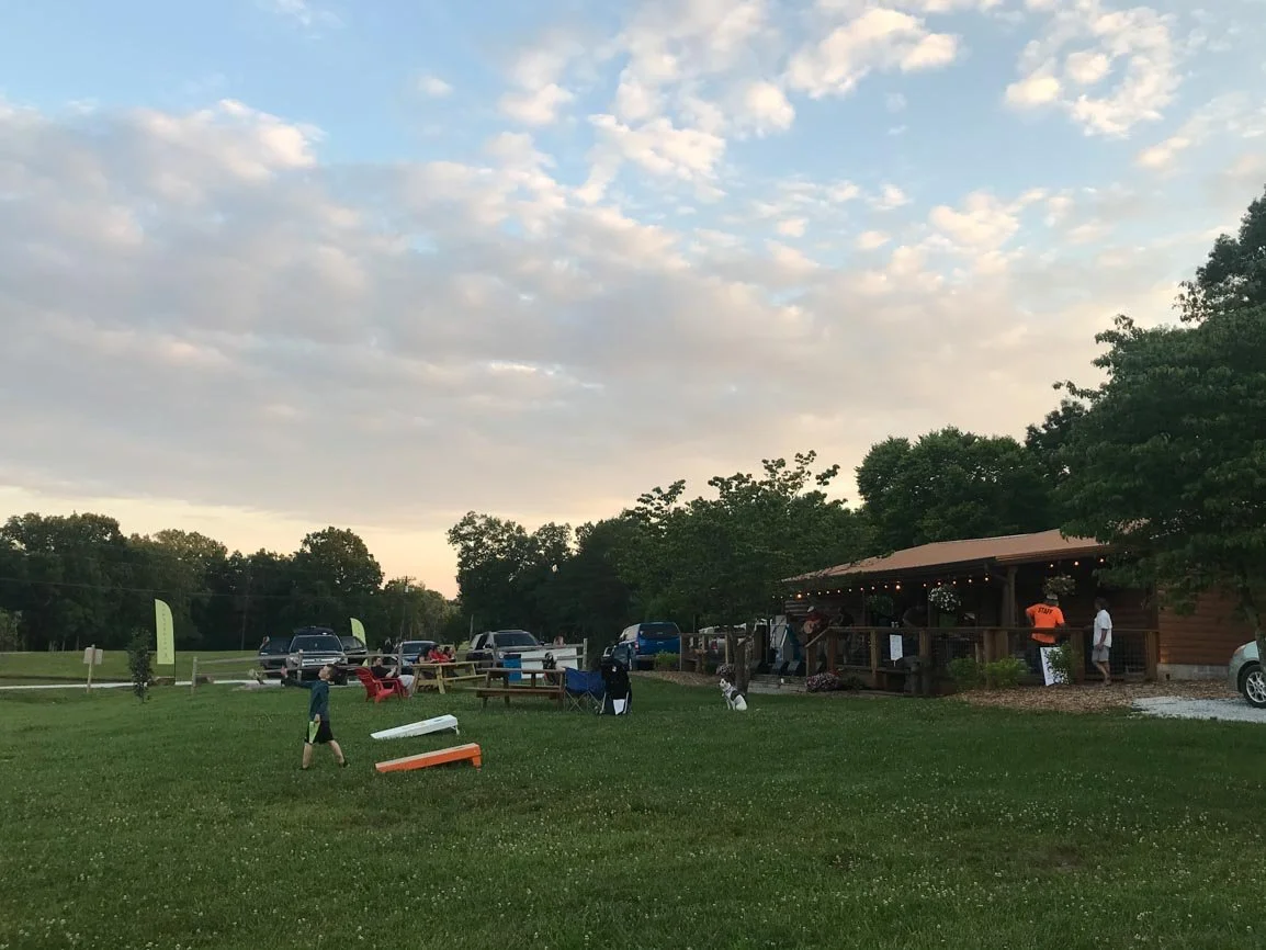 A park or outdoor event area during sunset with people walking, sitting in chairs, and socializing near a wooden building with string lights. Several cars are parked on the grass and a few trees surround the area.