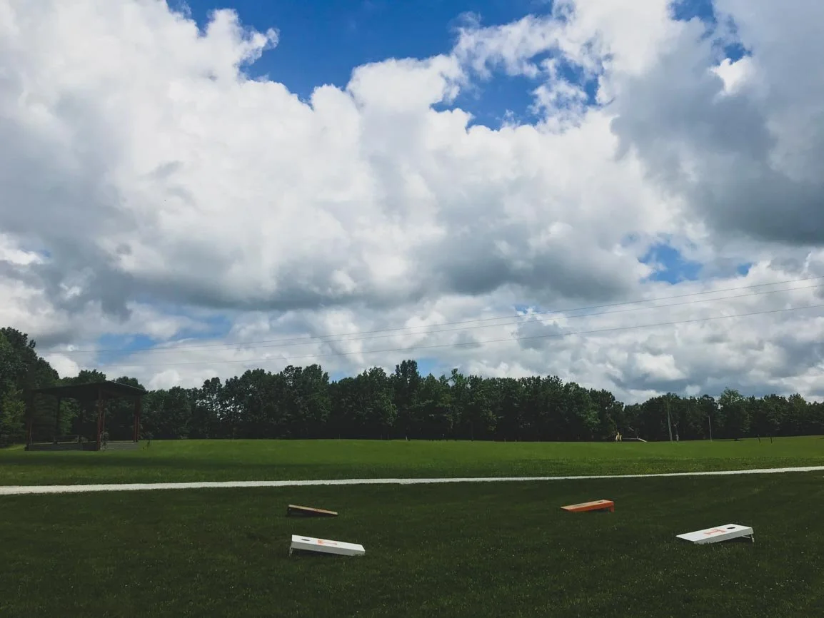 A grassy field with a dirt path running through it, surrounded by trees and under a cloudy sky. Cornhole boards are scattered on the grass in the foreground.