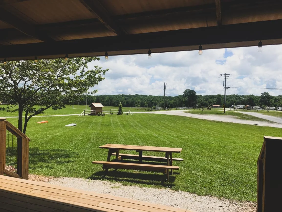 View from a covered porch looking out over a green lawn with a tree, a small shed, and a distant road with utility poles and vehicles under partly cloudy skies.