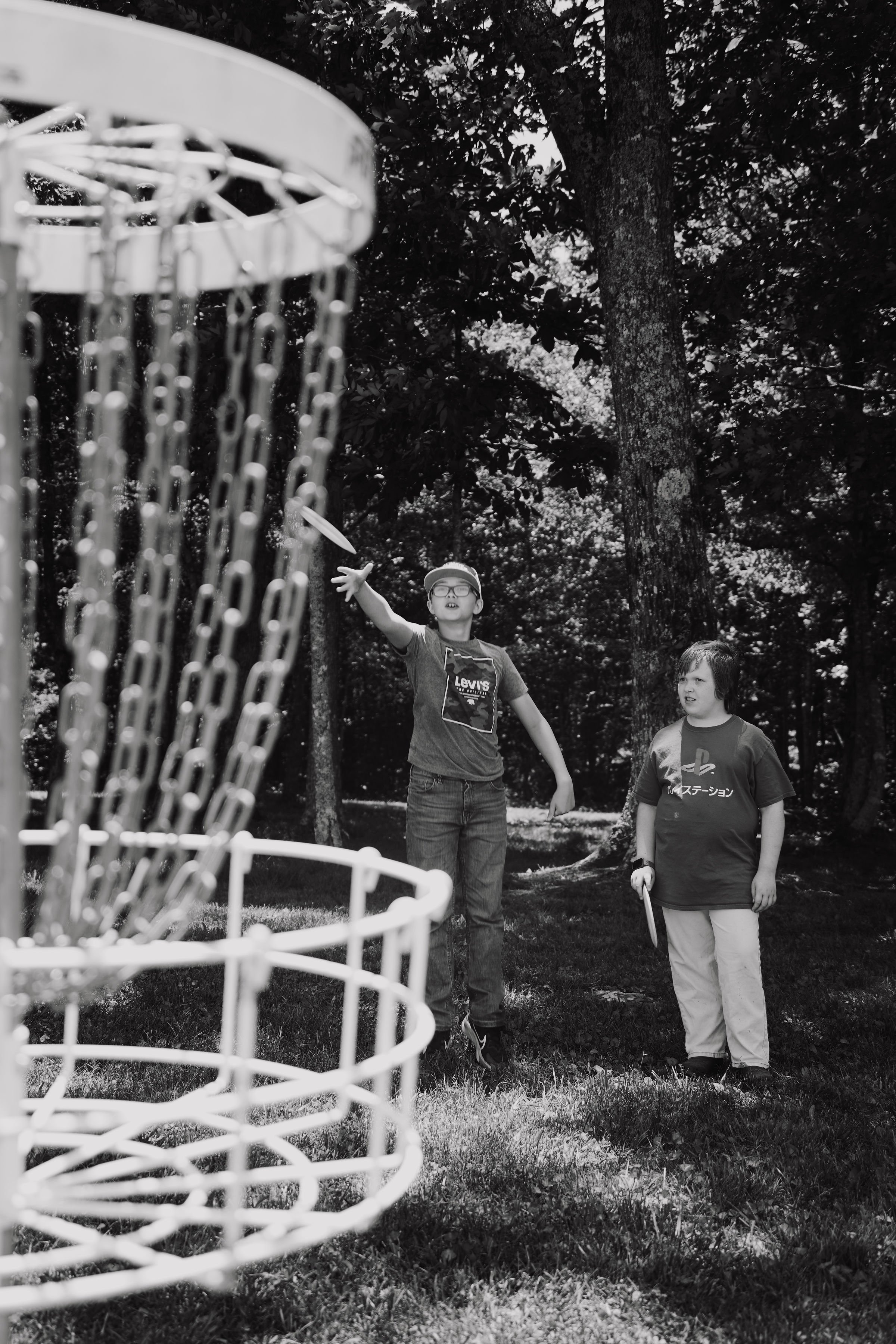 Two boys playing disc golf outdoors, one throwing a disc, with trees in the background.