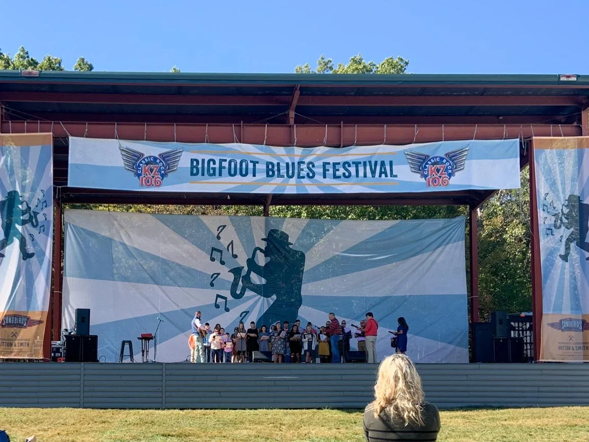 Stage with a large banner that reads "Bigfoot Blues Festival" and features a logo with wings and the number 106. A performer and a group of children are on stage, with a woman watching from the front. The background has a graphic of a saxophone player with musical notes, and the festival appears to be outdoors on a sunny day.