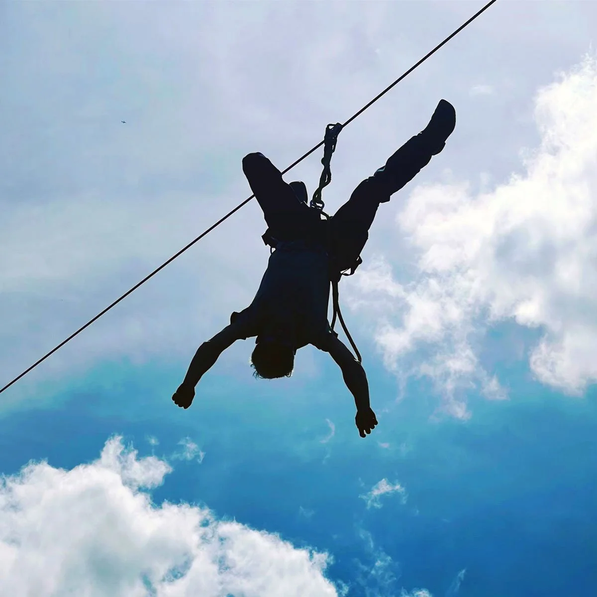 Person in harness, hanging upside down on a zip line in the sky with clouds in the background.