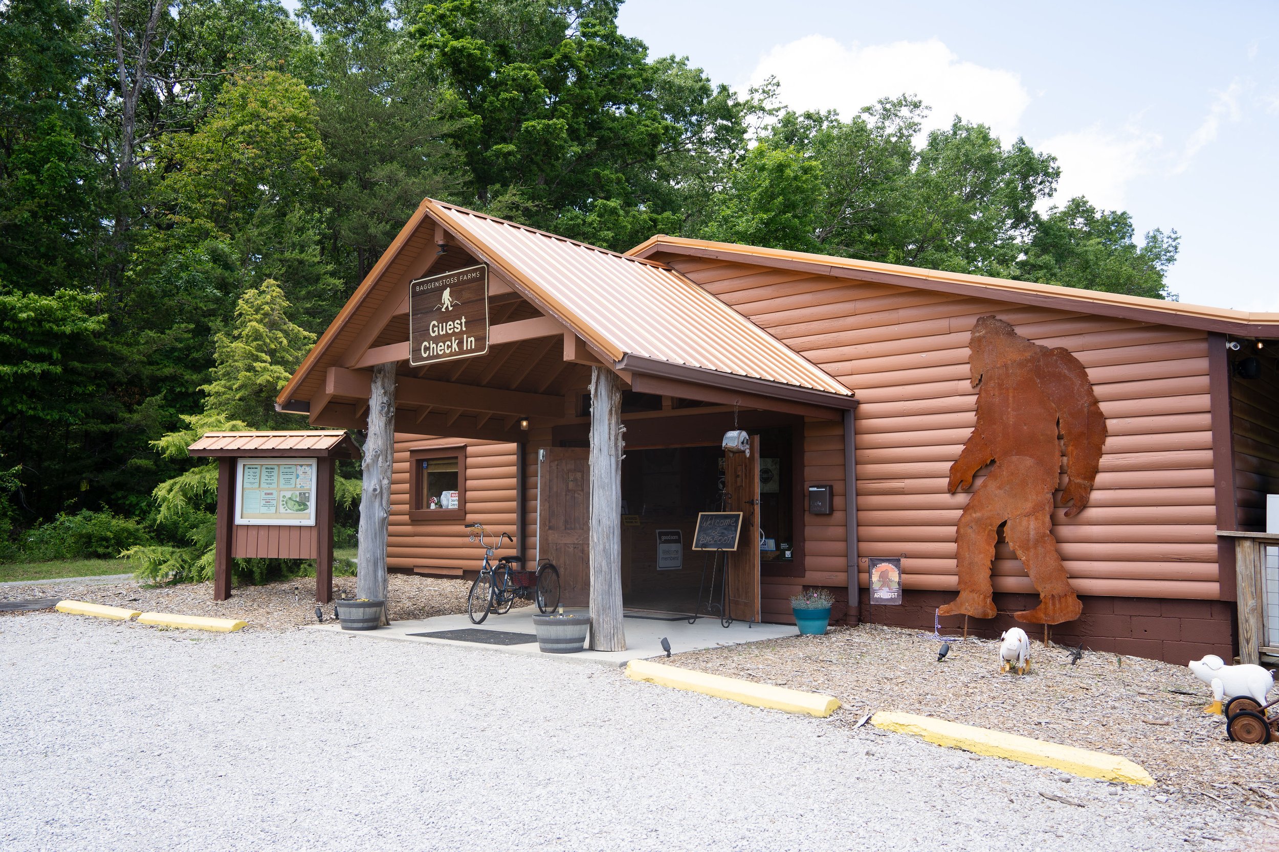 Rustic log building with sign that reads "Guest Check In," surrounded by trees. There is a 'Welcome' sign, a bicycle, planters, and decorative animal figures outside.