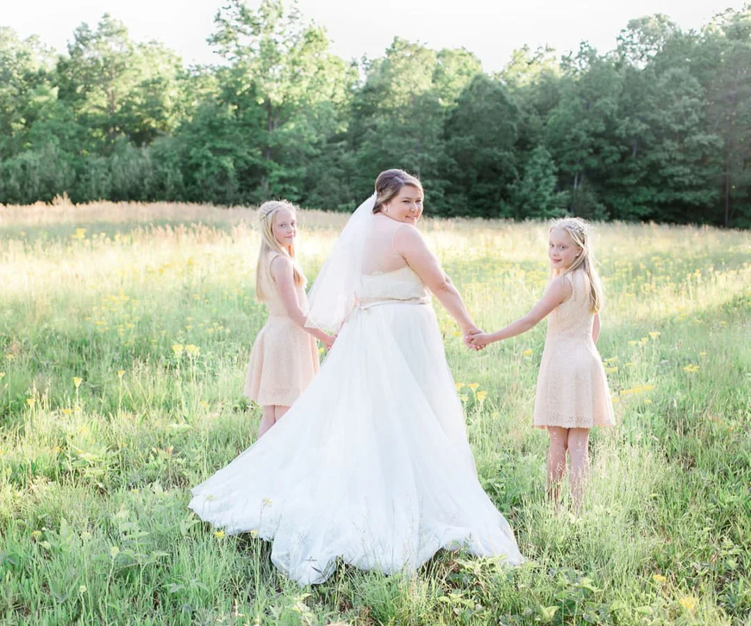 A bride in a white wedding dress holding hands with two young girls in pastel dresses in a green field with trees in the background.
