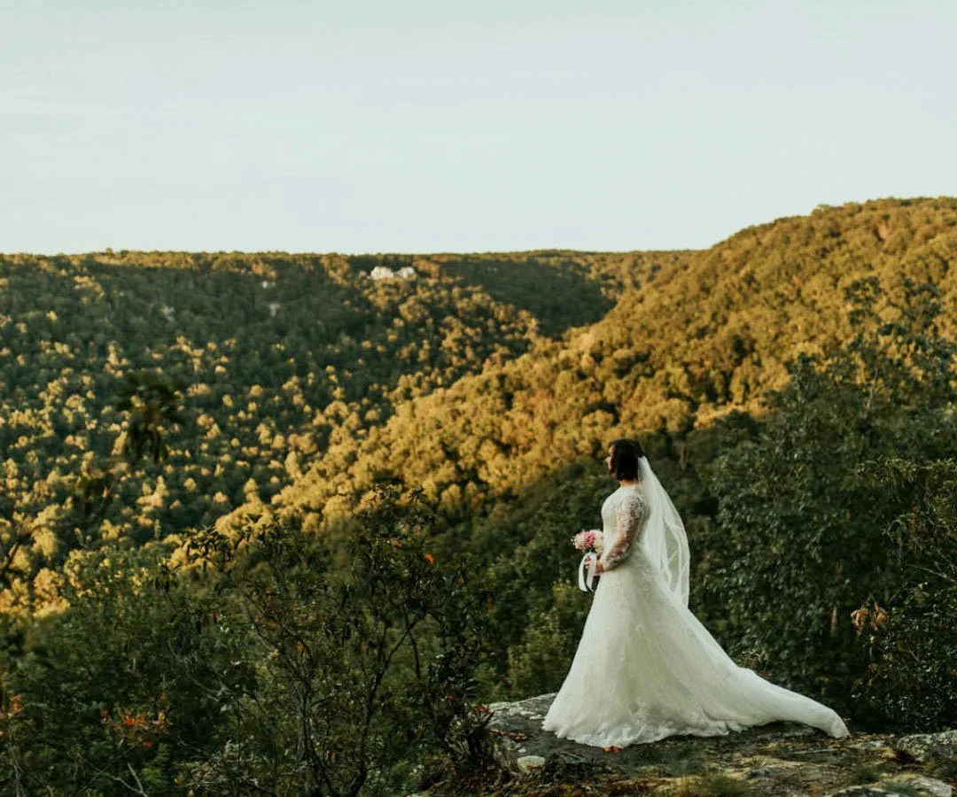 Bride in a white wedding dress holding a bouquet standing on a rocky ledge overlooking a lush green valley.