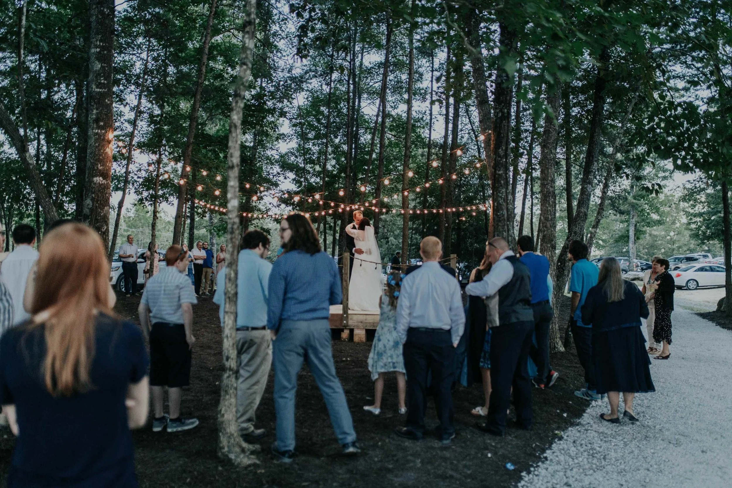 A wedding ceremony taking place outdoors in a wooded area with string lights overhead. The bride and groom are standing on a small platform, with guests gathered around.
