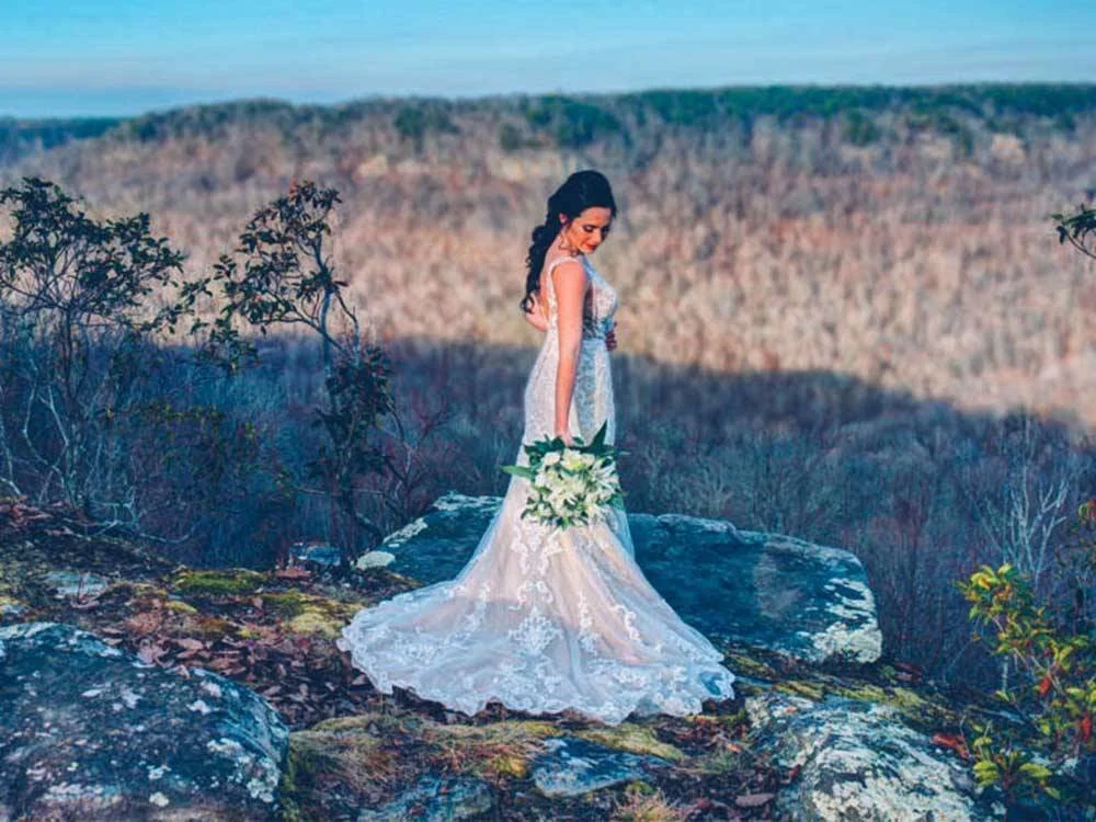 A woman in a white wedding dress holding a bouquet of white flowers, standing on a rocky outcrop with a forested landscape in the background.