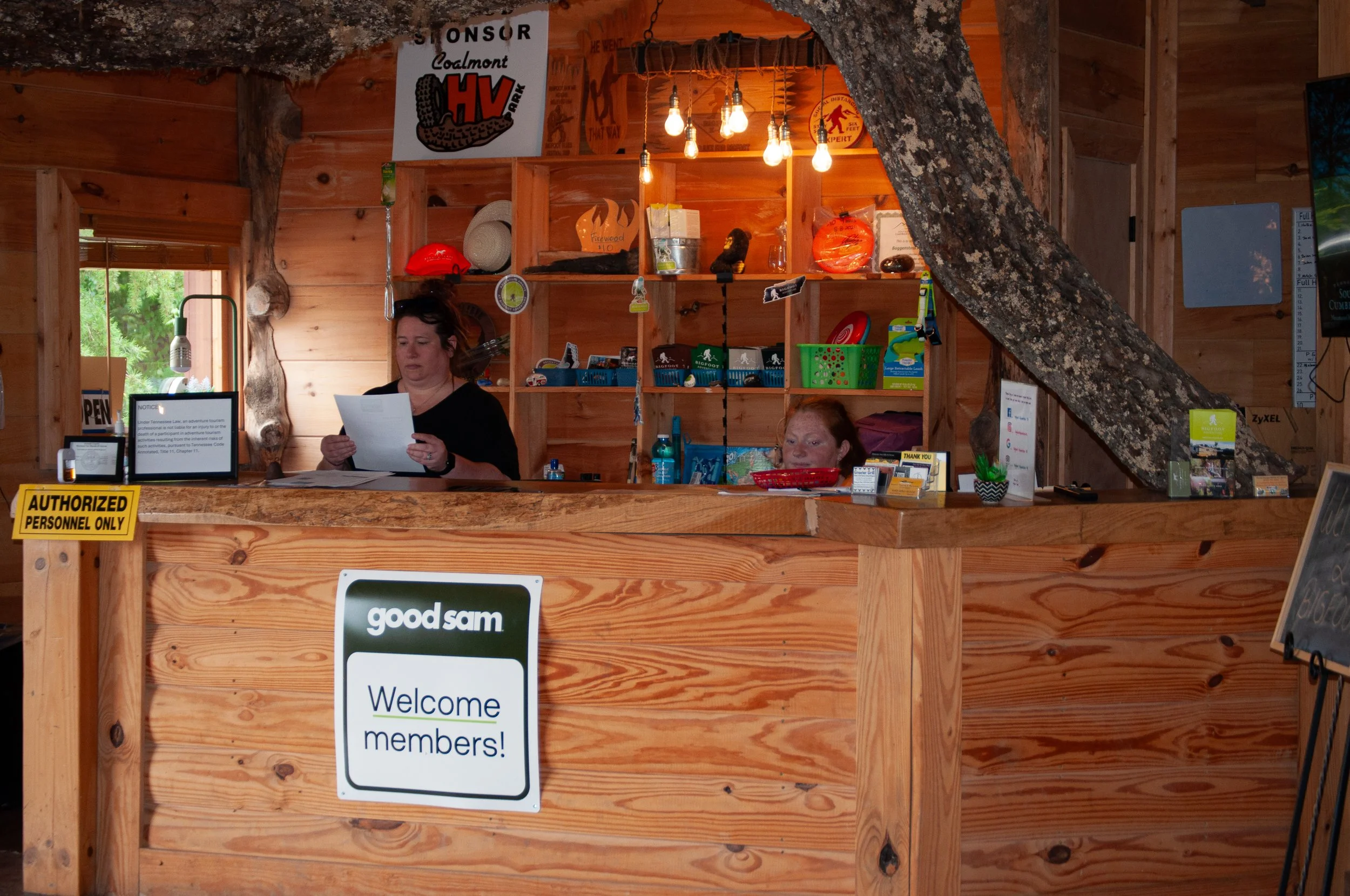Reception area with two women behind a wooden counter, one reading a paper and the other seated, in a rustic setting with wood walls, shelves, and various decorations including helmets, signs, and small items.