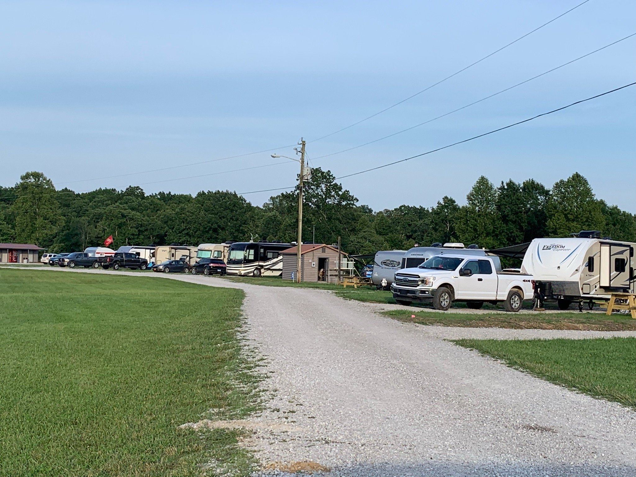 Camping area with RVs, trailers, and trucks parked along a gravel road, surrounded by green grass and trees in the background.