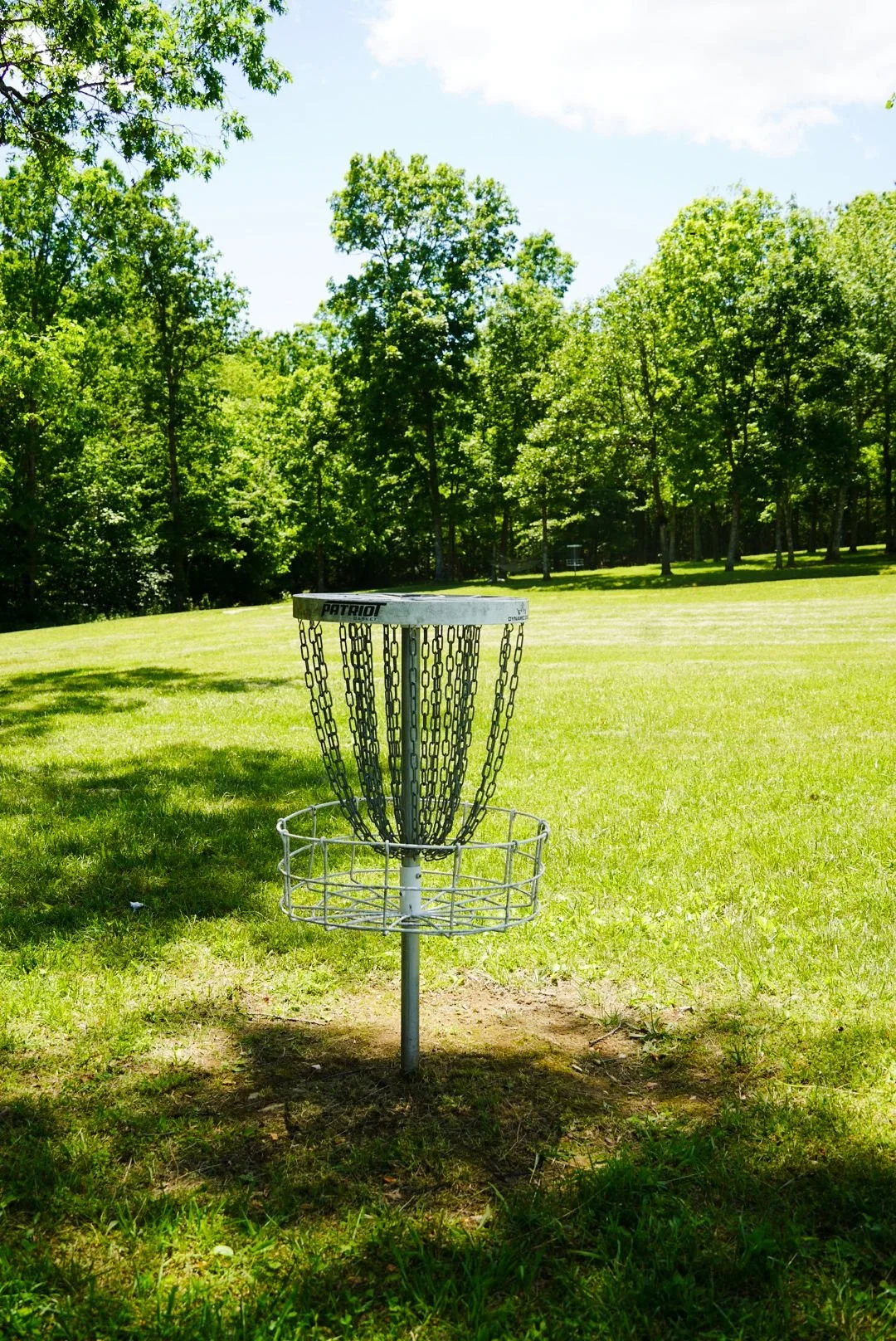 A disc golf basket with chains stands in the middle of a grassy park, surrounded by trees and under a bright, partly cloudy sky.
