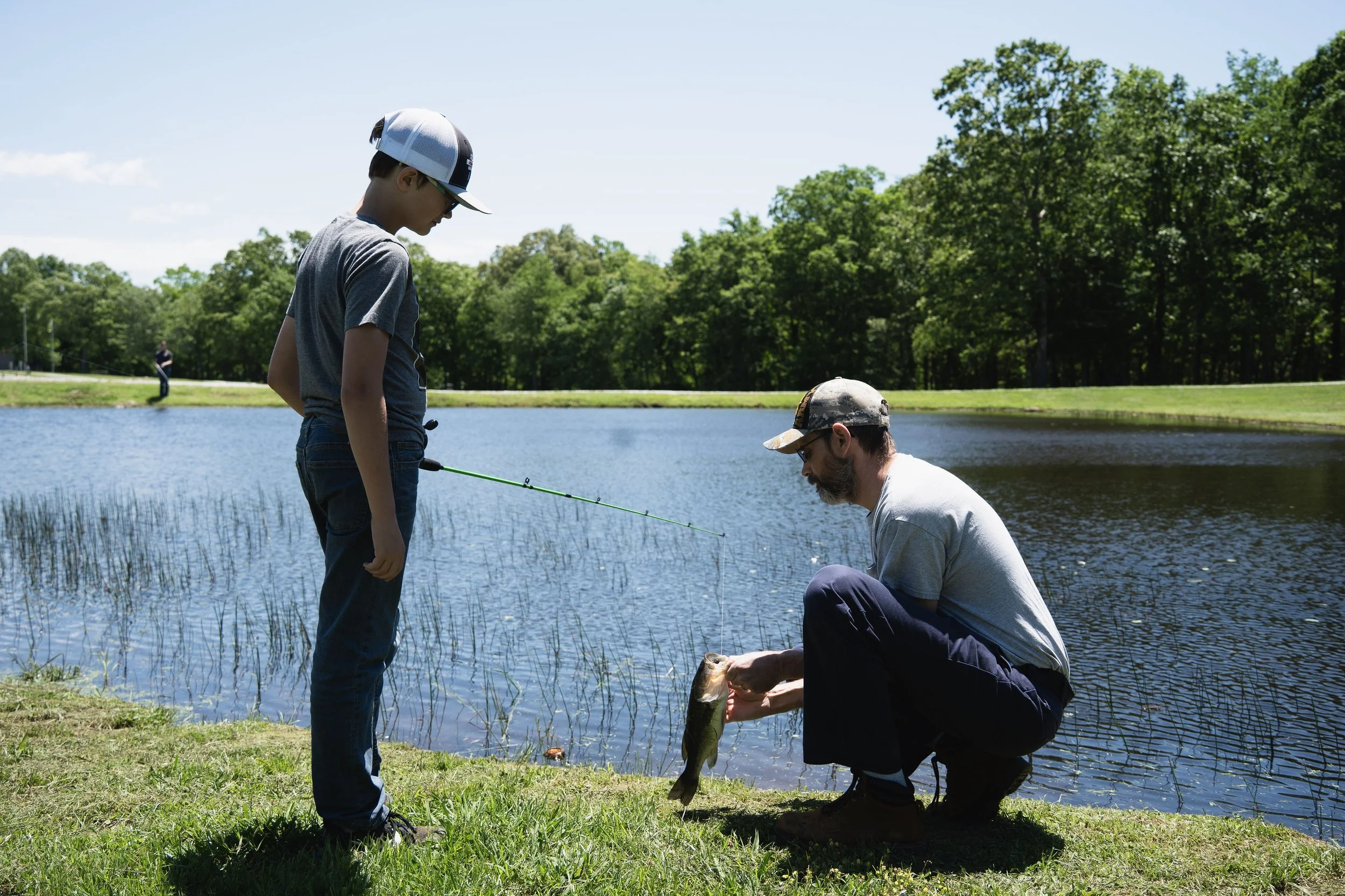 A man and a boy fishing by a lake surrounded by trees, with the man holding a caught fish.