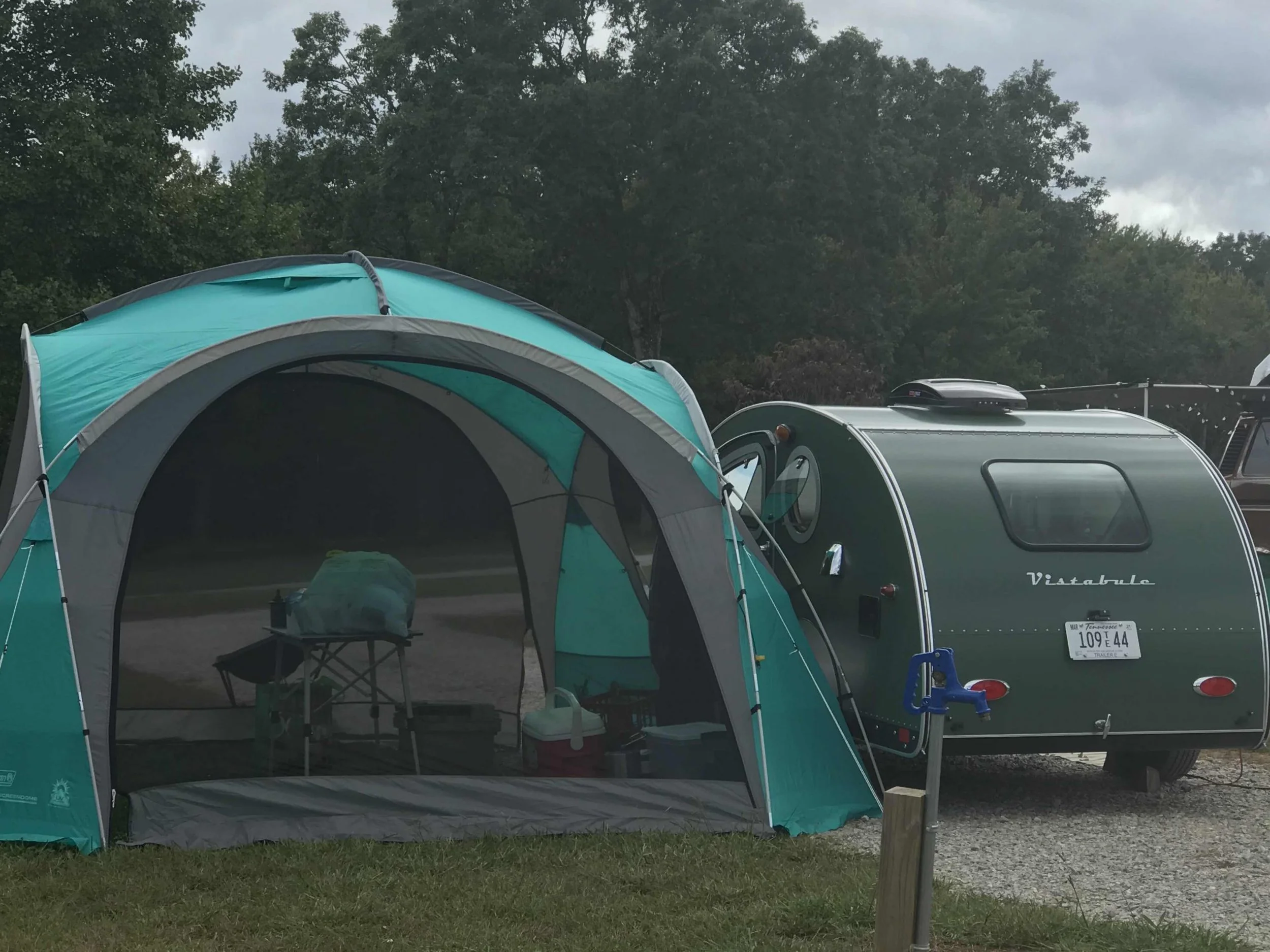 A teal and gray camping tent set up next to a small green travel trailer with a Tennessee license plate, with trees and cloudy sky in the background.