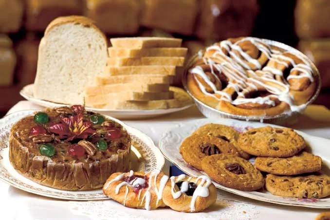 A variety of baked goods including a loaf of bread, sliced bread, a chocolate chip cookie cake, cinnamon rolls with icing, a donut with icing, and a plate of chocolate chip cookies.