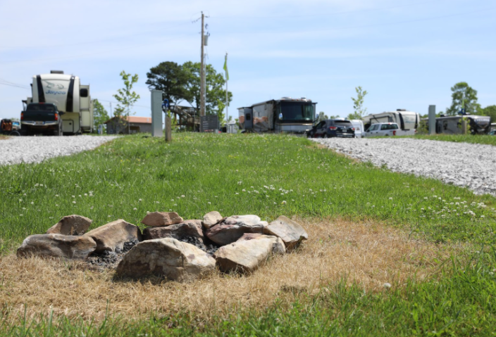 Park with a fire pit surrounded by rocks, grassy area, gravel pathway, and RVs or buses in the background on a partly cloudy day.