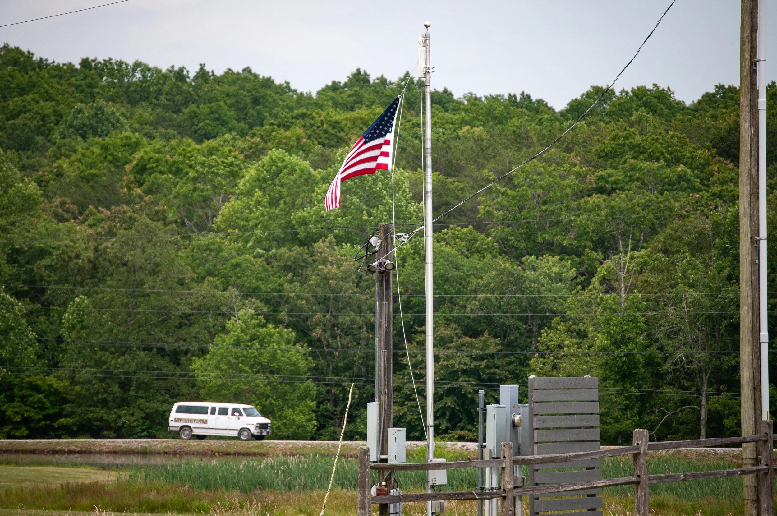 American flag flying atop a flagpole with lush green trees in the background and a white van in the distance.