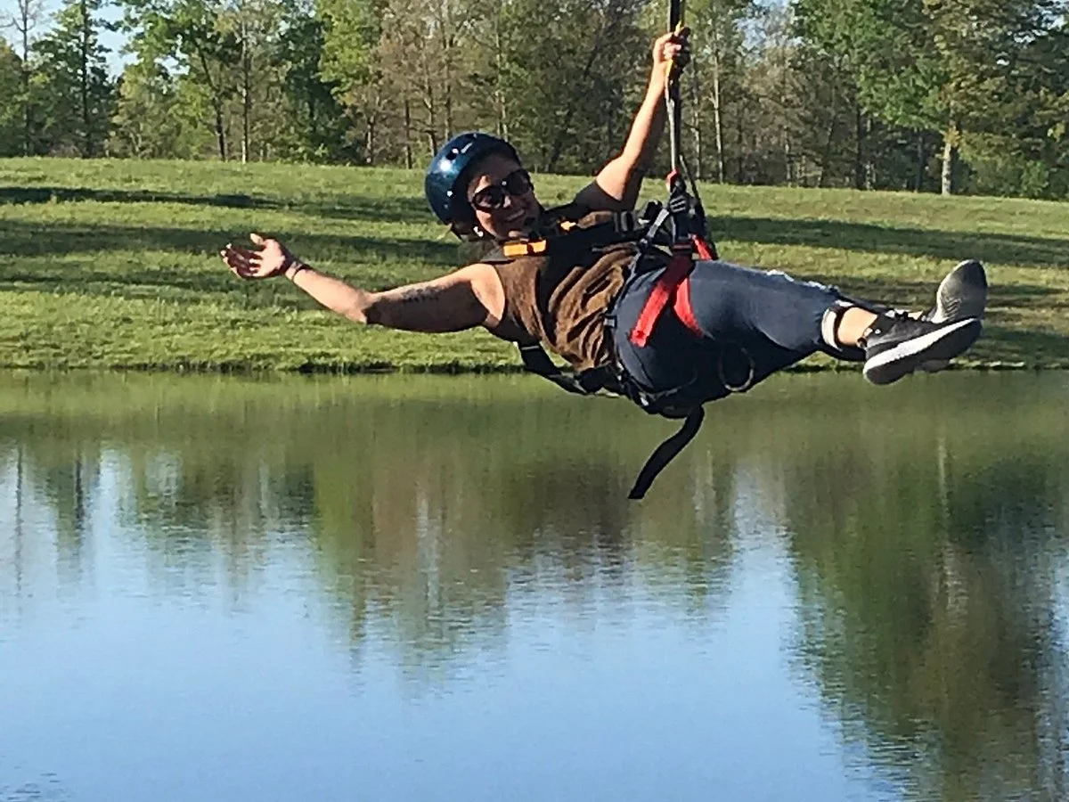 Person wearing a helmet and sunglasses riding a zip line over a lake, with their arms outstretched and smiling.