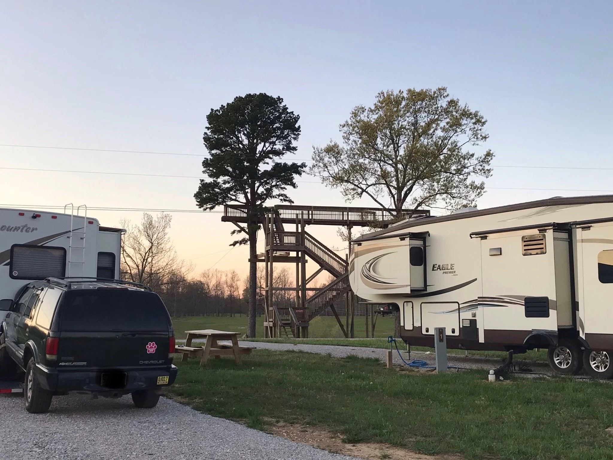 A campground with a black SUV, an RV, and a wooden treehouse on stilts connected by a deck, with two trees in the background during sunset.