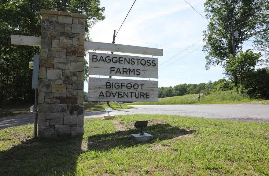 Sign at Baggentoss Farms advertising Bigfoot Adventure situated near a rural road with trees and open fields in the background.