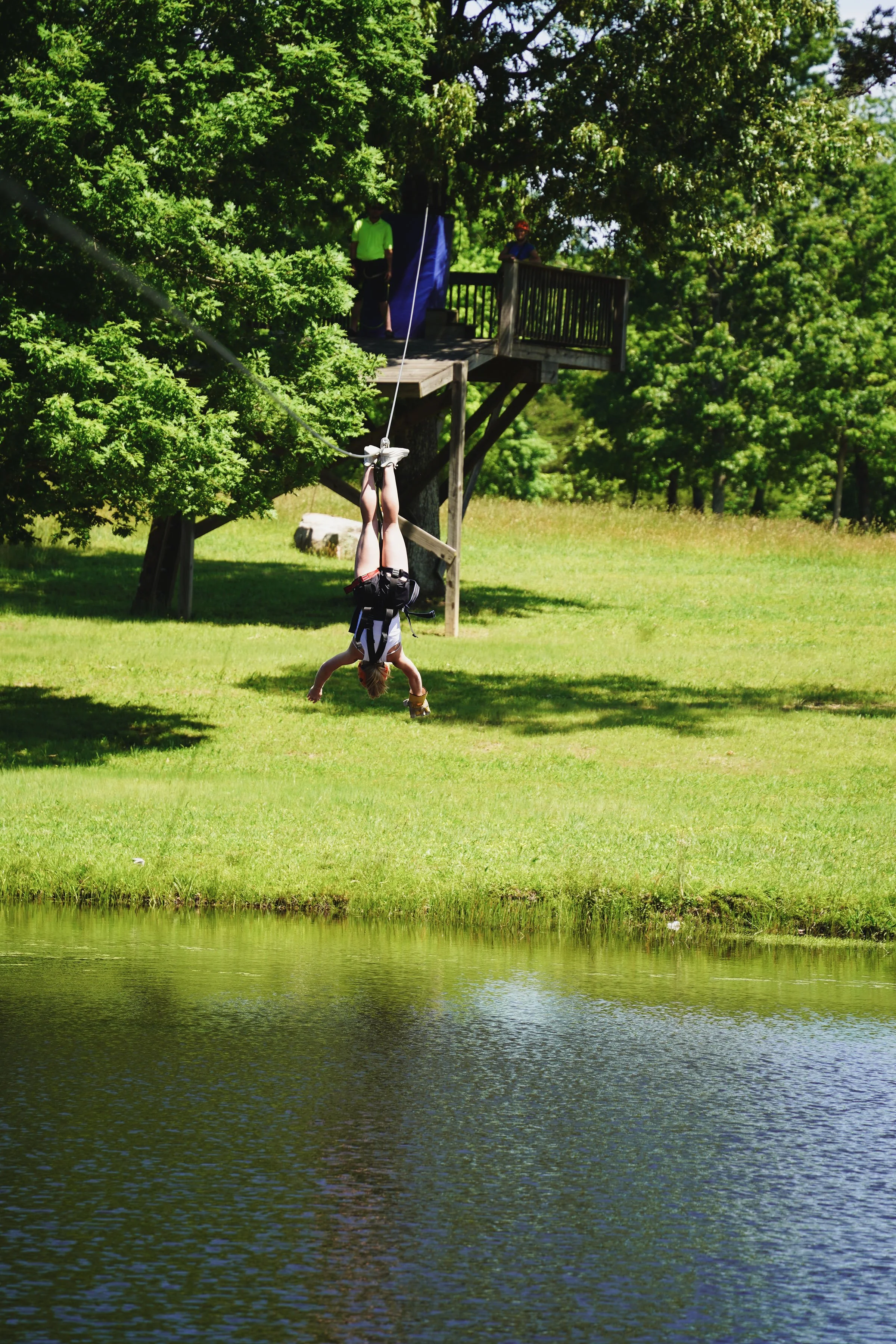 Person hanging upside down on a zip line over a body of water with trees and a wooden platform in the background.