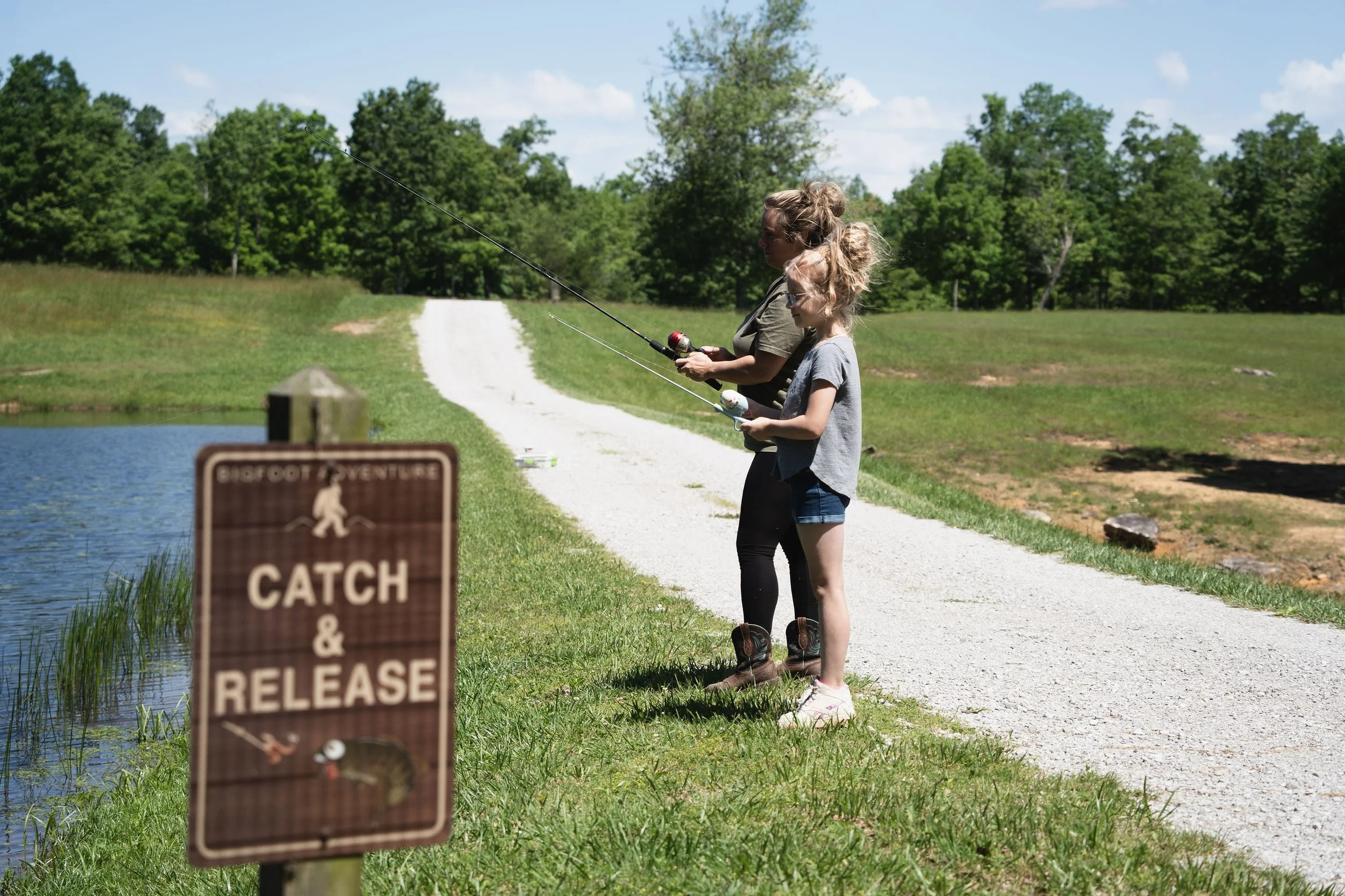 A woman and a girl fishing by a small lake in a park, with a brown sign in the foreground that reads "Catch & Release."