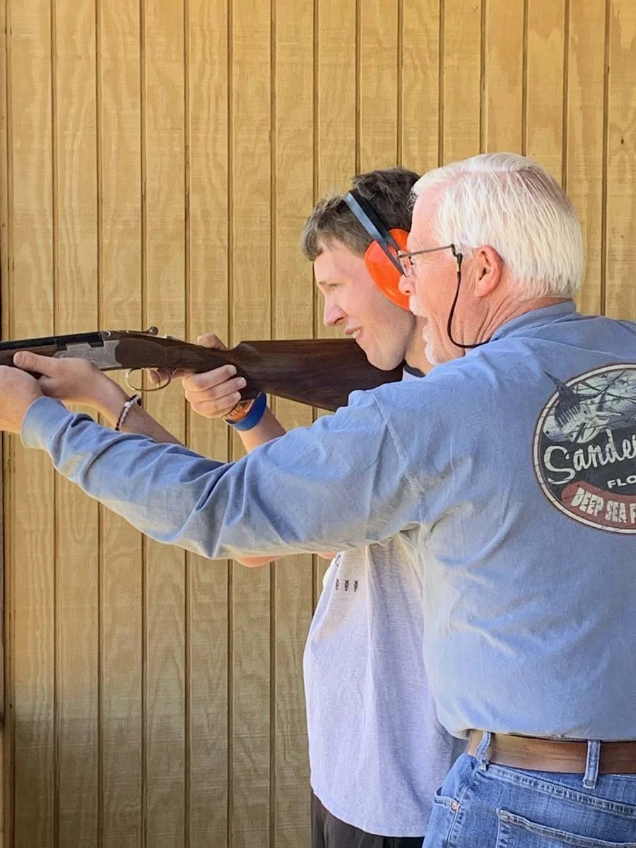 An elderly man teaches a young man how to shoot a rifle at a shooting range, with both aiming and focusing on the target.