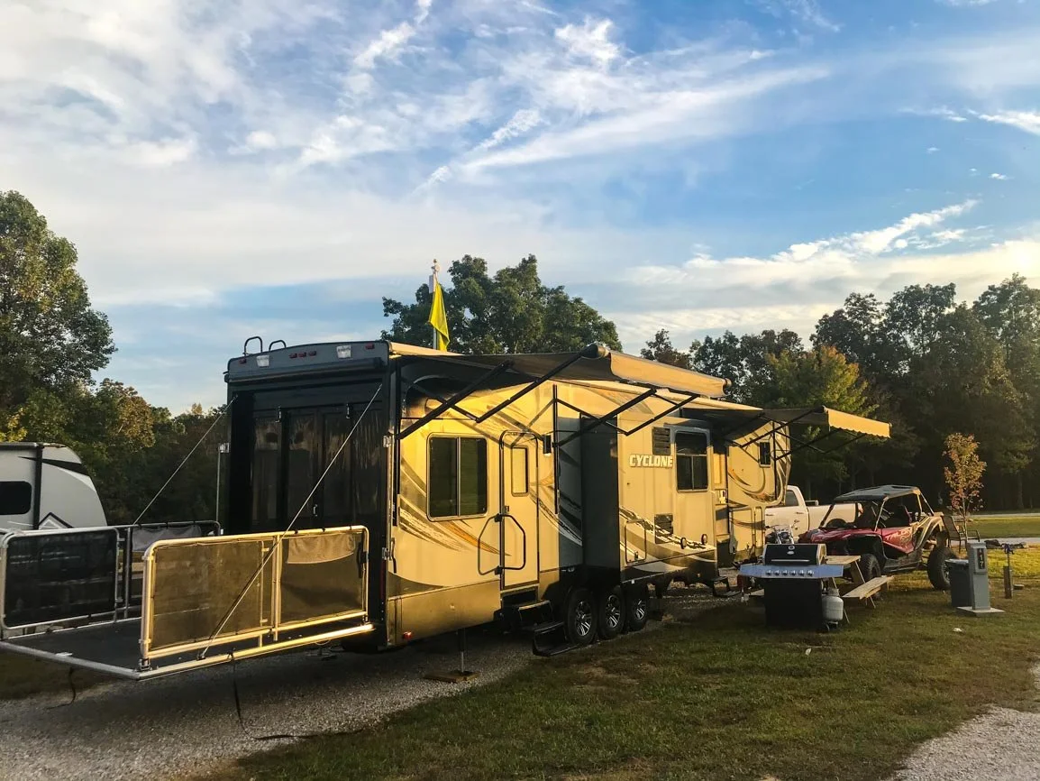 A yellow and black camper trailer labeled 'Cyclone' parked outdoors next to an ATV with a side-by-side vehicle, barbecue grill, and trash bin on grassy terrain with trees and a partly cloudy sky in the background.